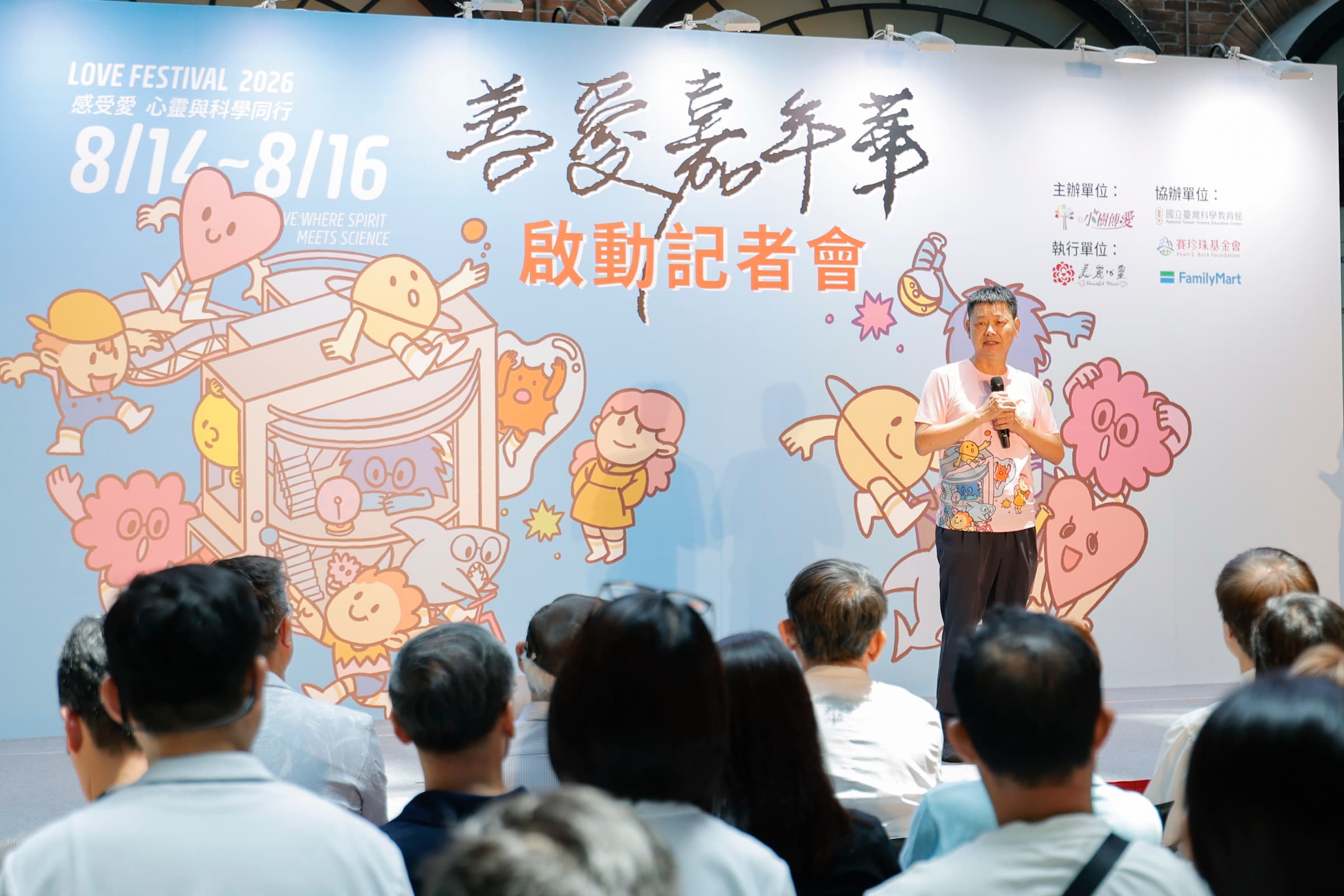 身著小樹傳愛粉色 T 恤的主辦代表於舞台手持麥克風致詞，身後為 2026 善愛嘉年華主視覺背板，台下媒體與來賓就座聆聽