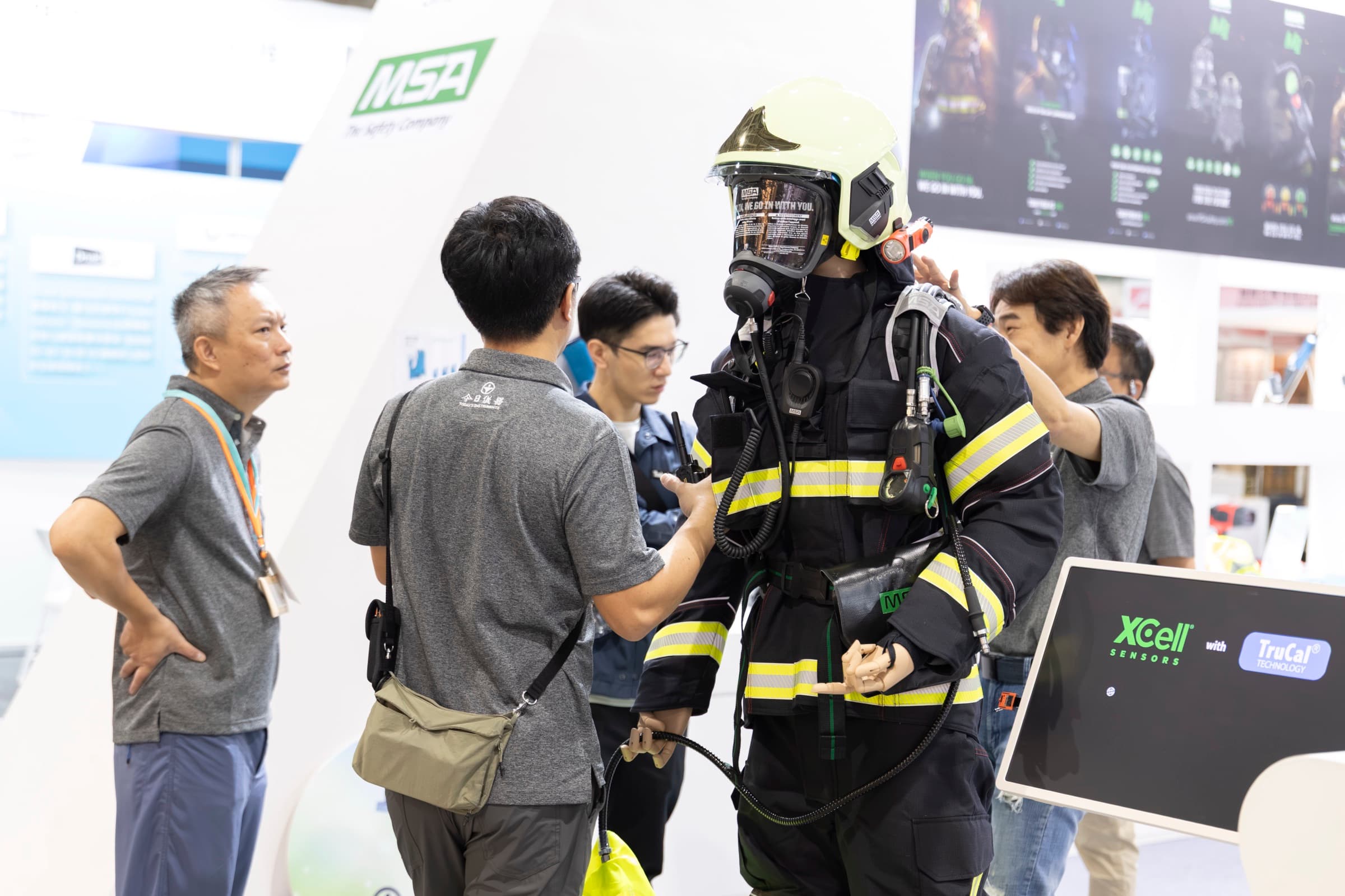 Secutech 展場消防安全展區，參展商為體驗者穿戴整套消防員空氣呼吸器與反光防護戰鬥服，旁有觀眾近距圍觀