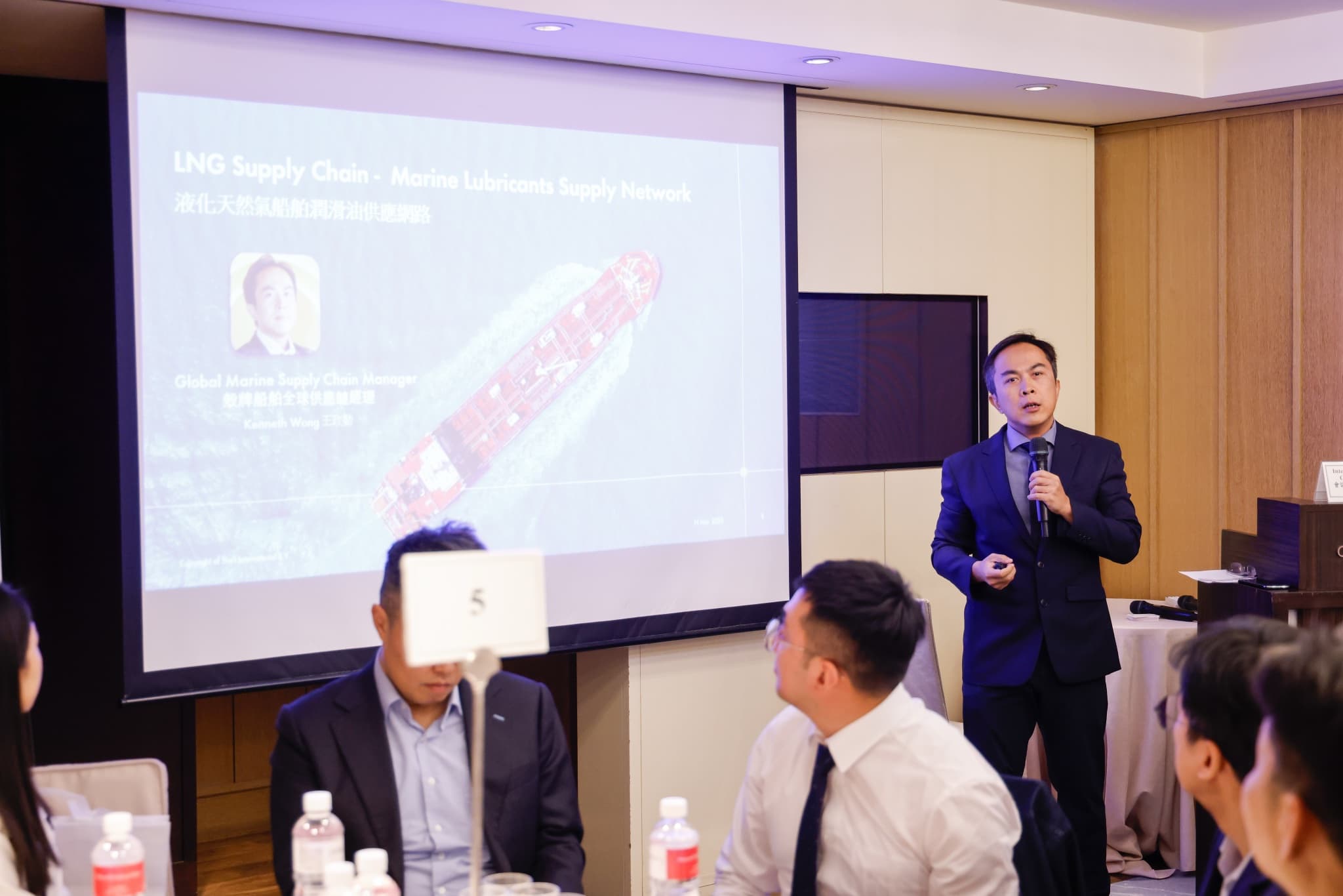 講者於台前持麥克風簡報，投影幕顯示 LNG Supply Chain — Marine Lubricants Supply Network 液化天然氣船舶潤滑油供應網路標題與貨櫃船空拍圖，前排與會者抬頭聆聽
