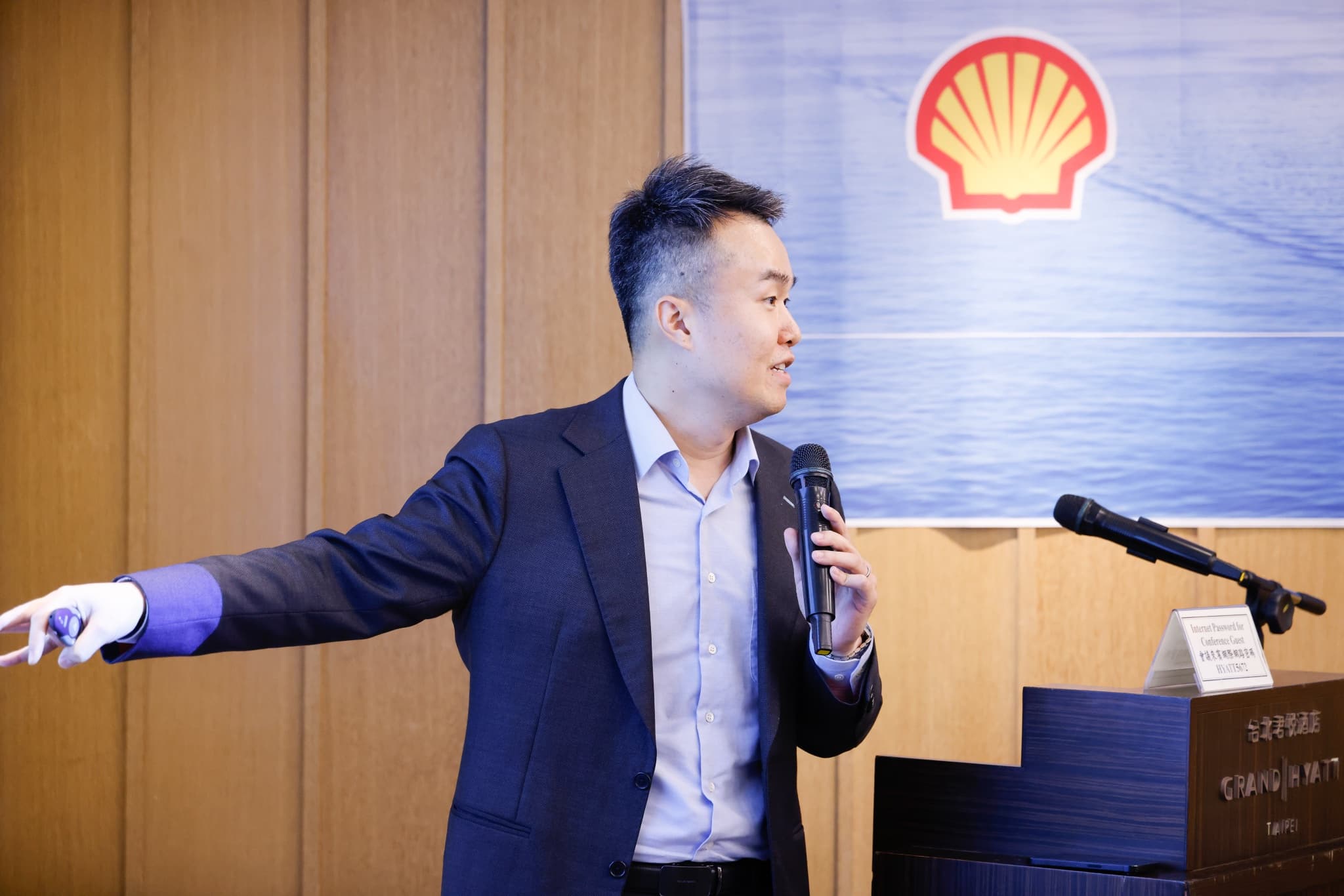 講者持麥克風側身伸手指向簡報方向，身後懸掛印有大型 Shell 貝殼標誌的藍色海面背板旗