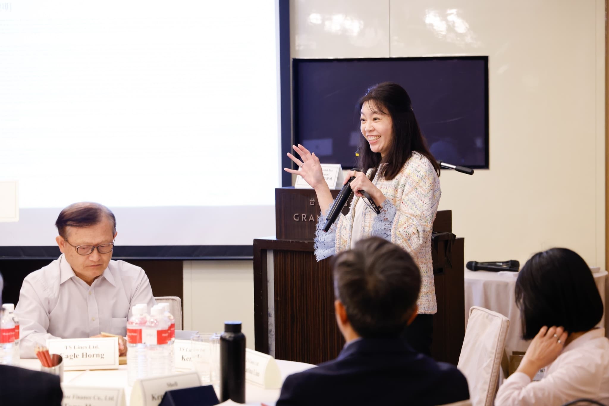 女性講者持麥克風站在講台旁微笑向與會者說明並舉手示意，前排桌上立有 TS Lines Ltd. 與 Shell 桌牌