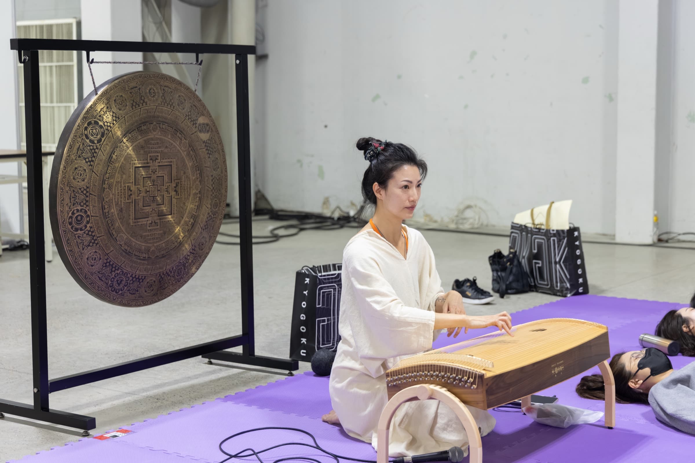 睡眠療癒展音療體驗區，身著白衣的音療師跪坐彈奏古箏，一旁架著大型銅鑼（頌缽），參與者戴眼罩躺臥於紫色地墊上放鬆