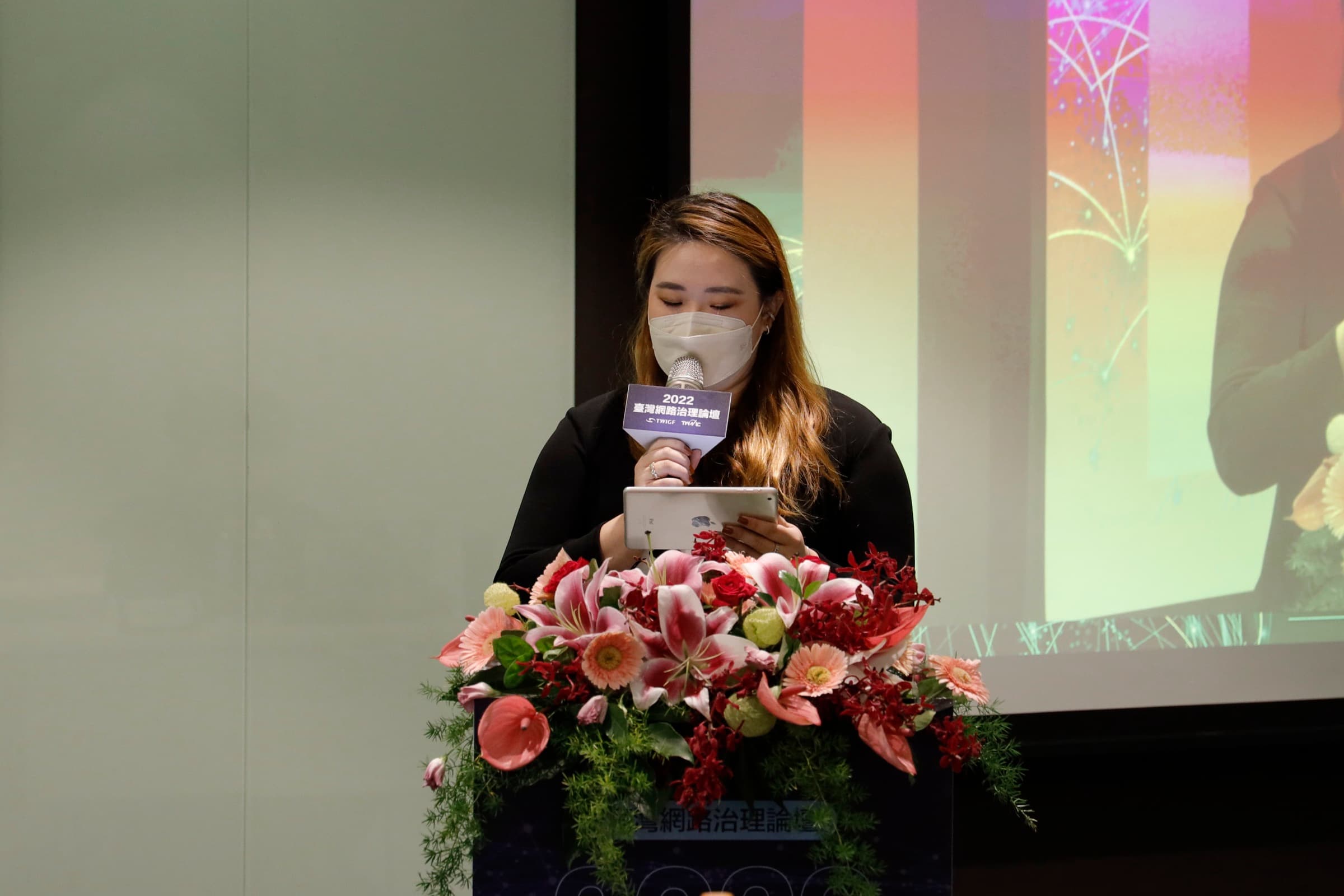 一位女性主持人於擺放花藝的講台後手持麥克風與平板主持，麥克風牌寫著『2022 臺灣網路治理論壇』