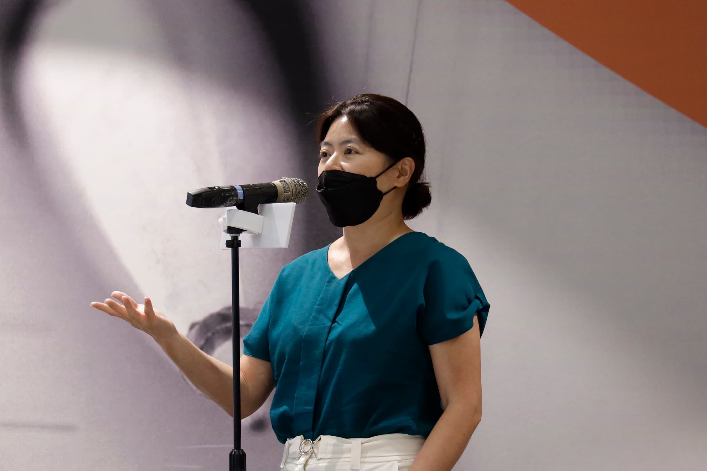 一位身著藍綠色上衣的女性與會者於落地麥克風前起身提問、手勢說明