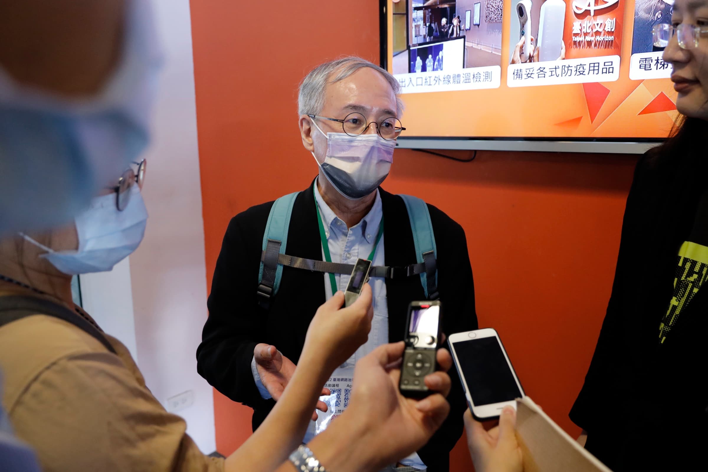 一位配戴識別證的與會代表被多支手機與錄音筆圍繞受訪，背景螢幕顯示臺北文創防疫資訊