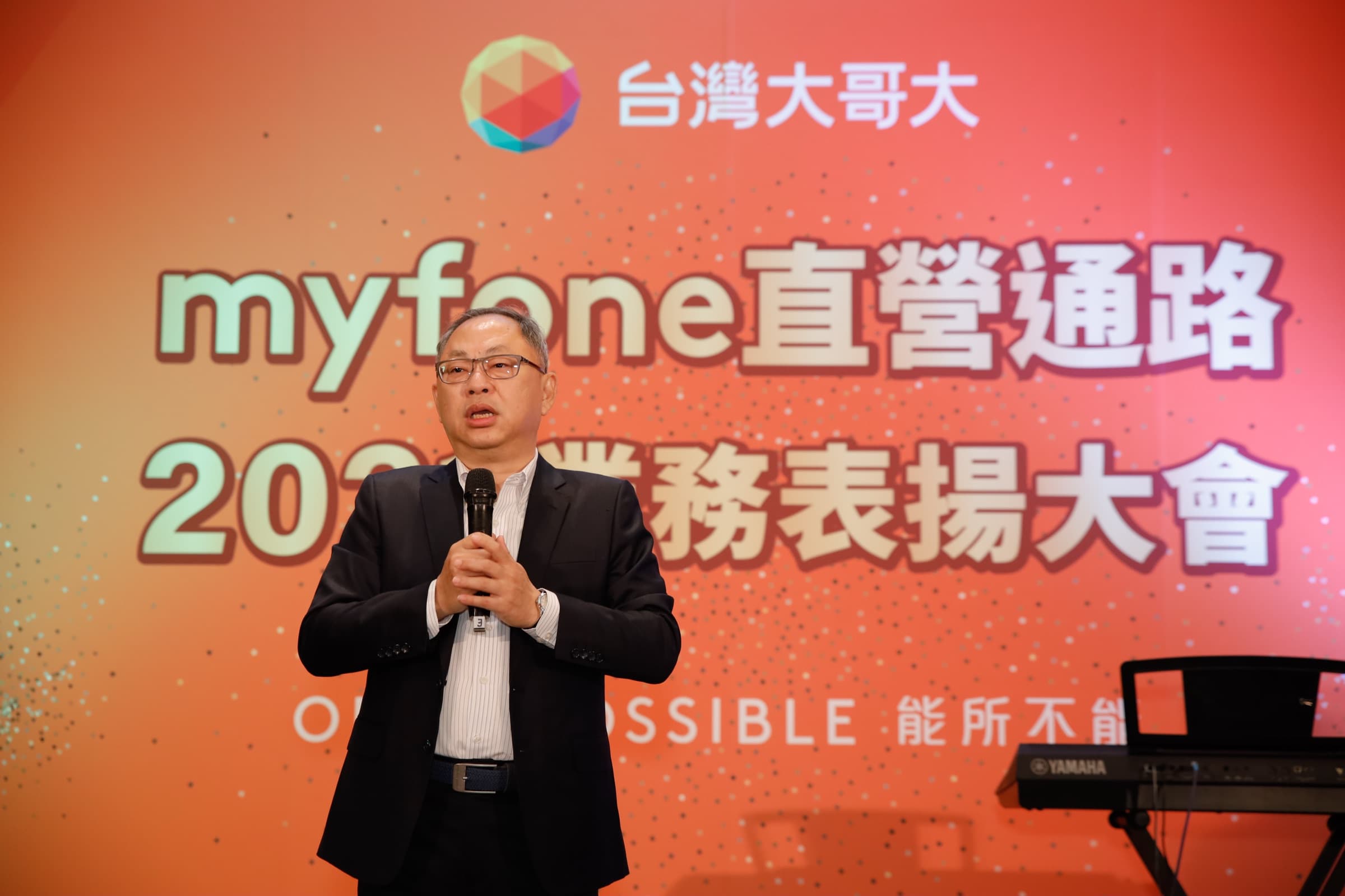 一位西裝男士手持麥克風於『myfone 直營通路 2021 業務表揚大會』背板前致詞