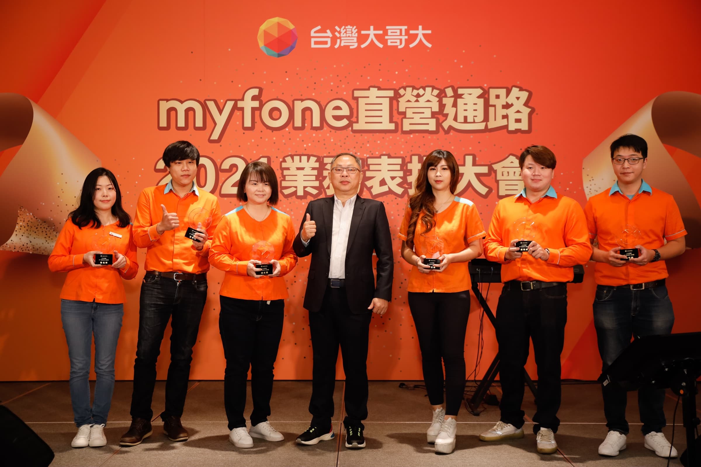 六位身著橘色制服的績優同仁各持玻璃獎座，與中央一位西裝男士於主視覺背板前比讚合影