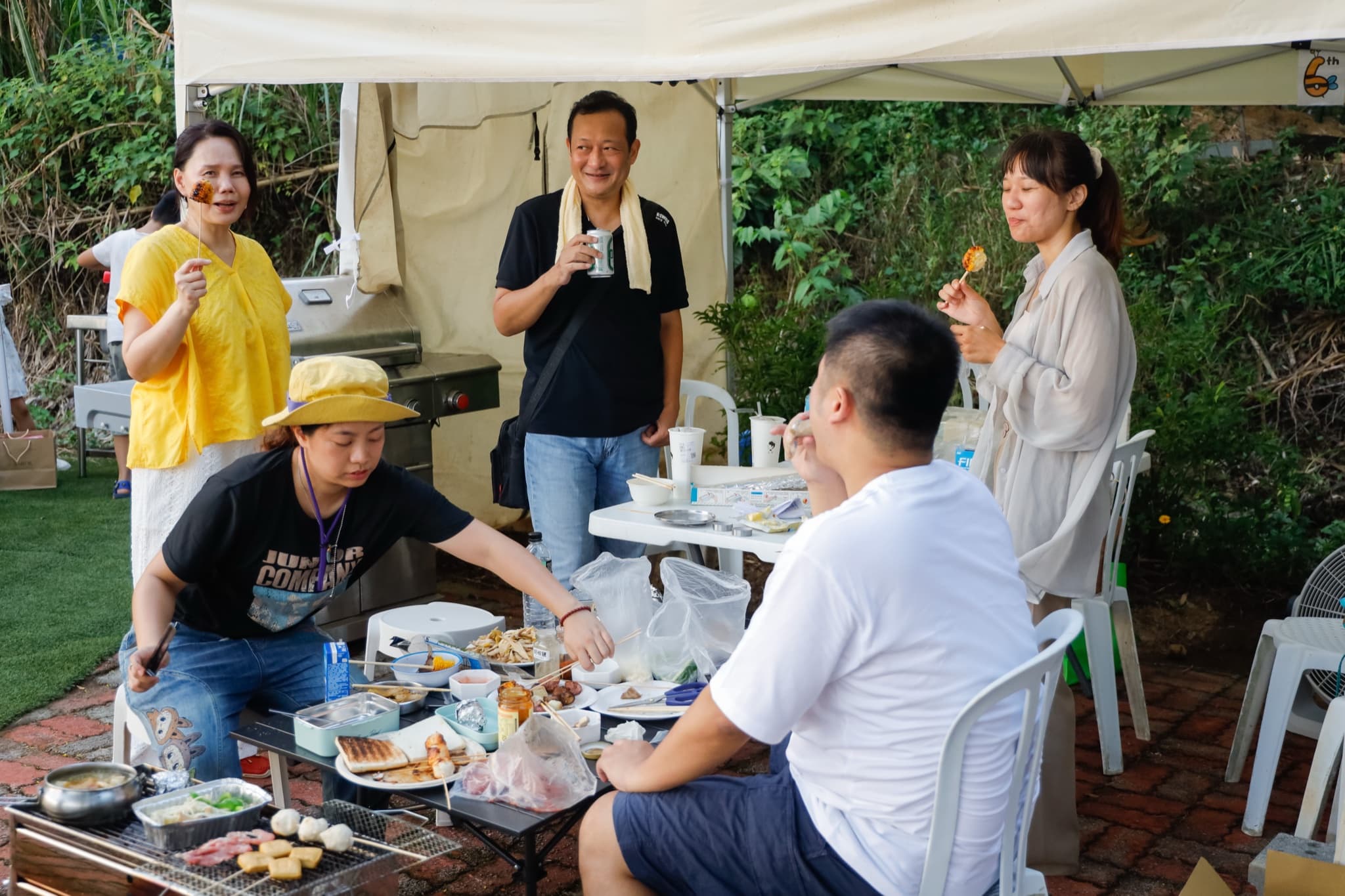 五位成員在帳篷邊圍著矮桌烤肉聊天,穿黃衣的女士舉著烤串,站立的男士拿著啤酒微笑,桌上擺滿食材與烤盤