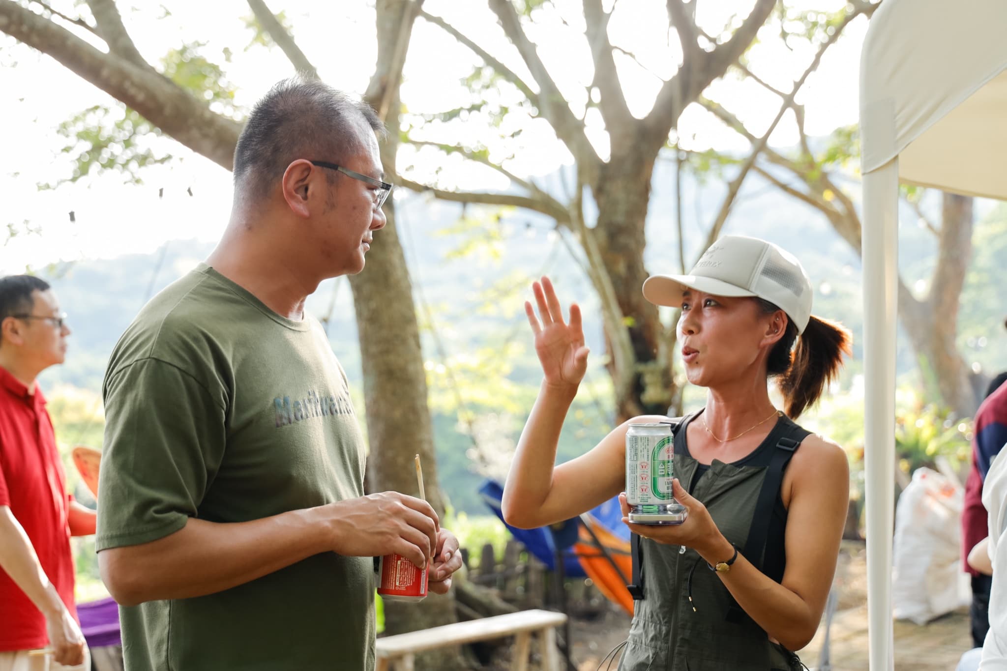 兩位成員在樹下拿著啤酒罐交談,右側戴帽的女士舉手比劃說話,背景是逆光的山景與帳篷一角