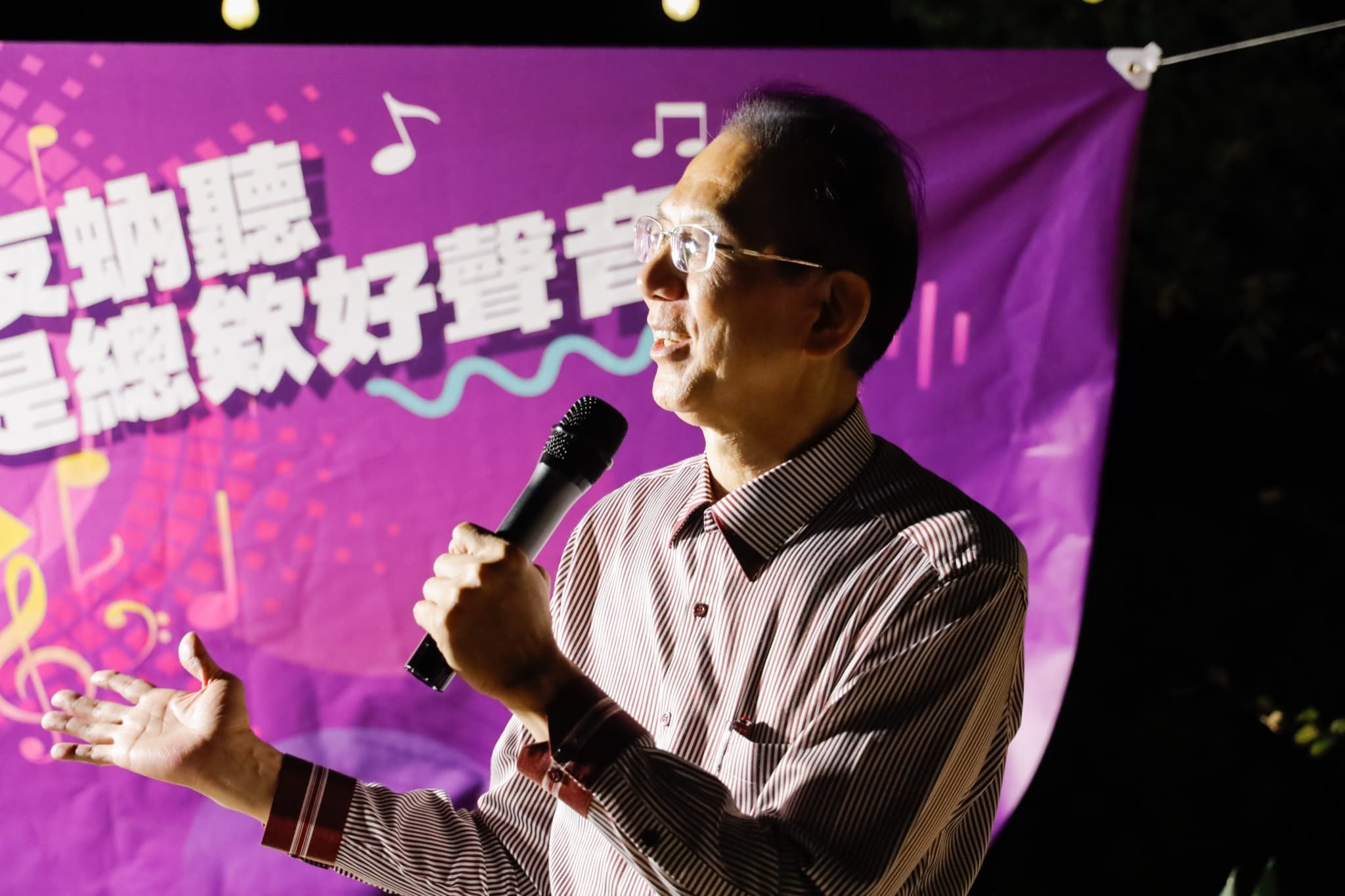 一位資深成員在紫色音樂主題背板前拿麥克風演唱,另一手張開比劃,背板印有台語標語與音符圖案