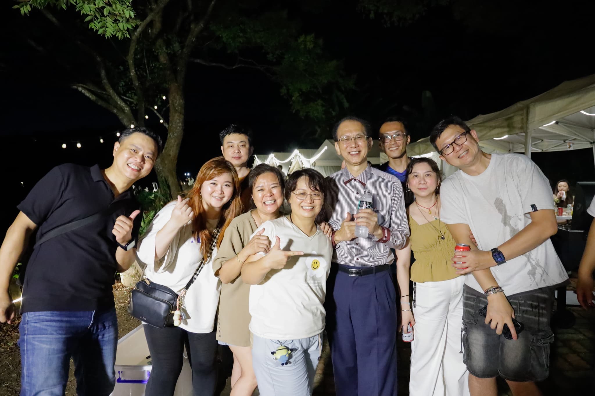 九位成員在夜色中的帳篷與樹下合影,比讚與手勢,人人面帶笑容,遠處帳篷燈串亮著