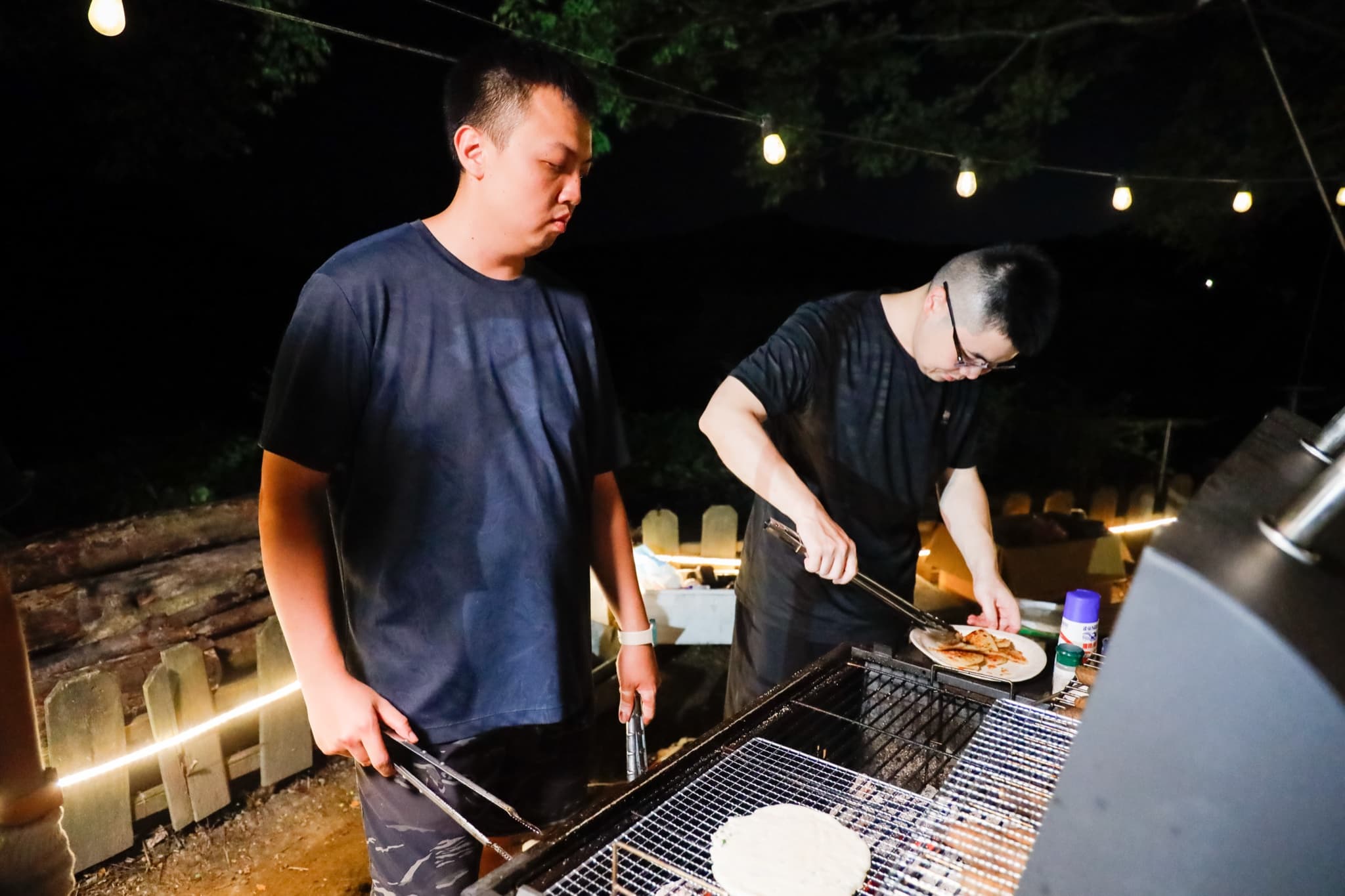 夜間串燈下兩位成員在烤爐前作業,一位持長夾翻烤、一位裝盤,木柵欄纏著發光燈條