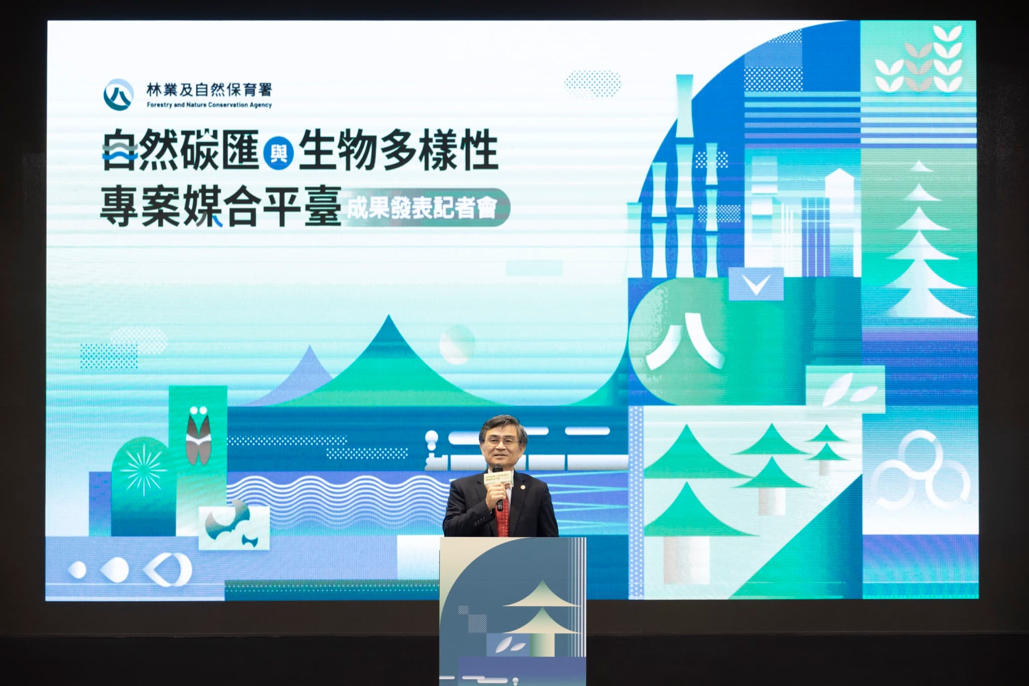 長官於講台致詞的舞台全景,LED 螢幕主視覺完整入鏡,下方為深色舞台與印有活動圖案的講台。