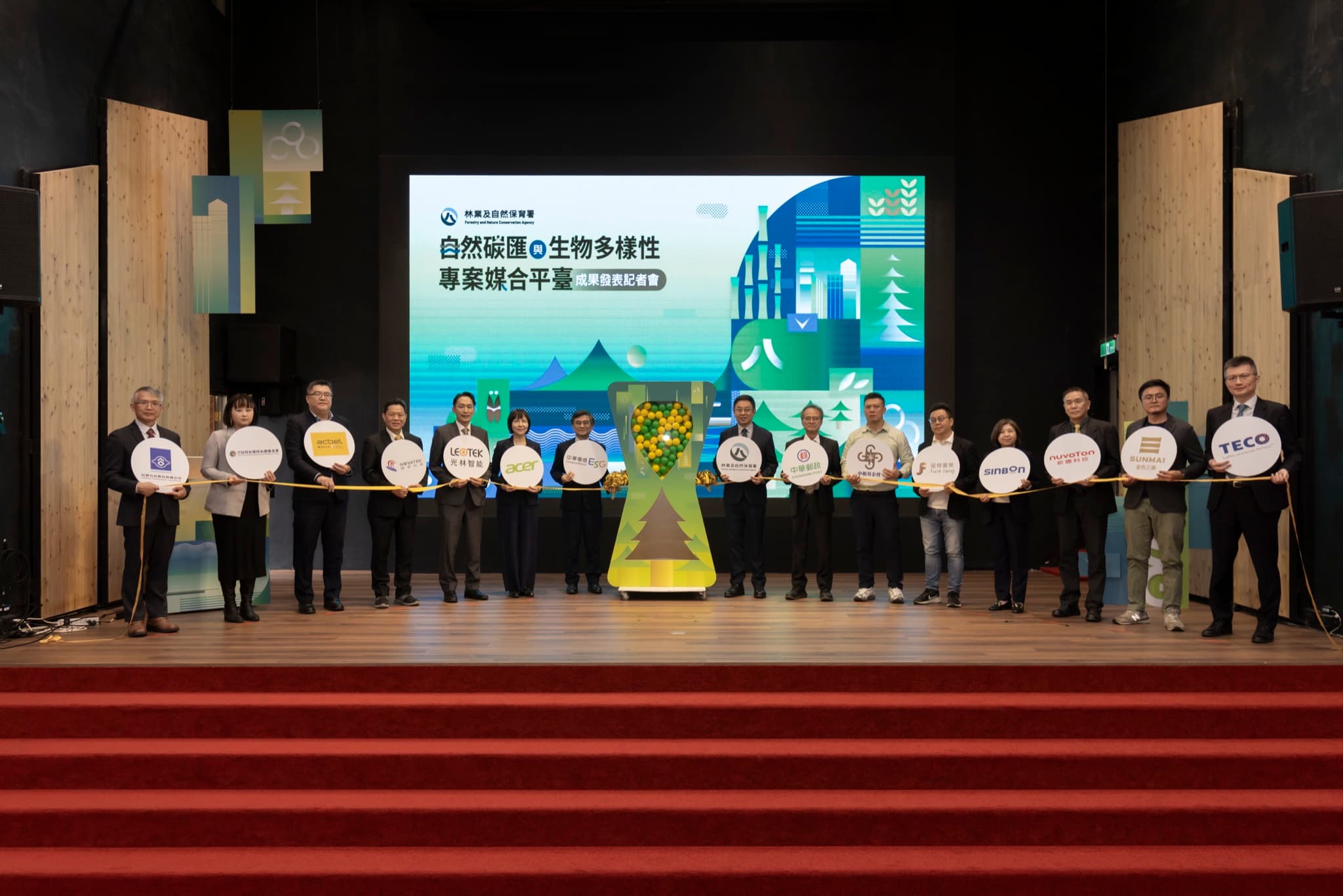 十六位長官與企業代表在舞台上共同拉起金色繩索,中央為裝滿綠黃色球的沙漏型裝置,背後為 LED 主視覺。