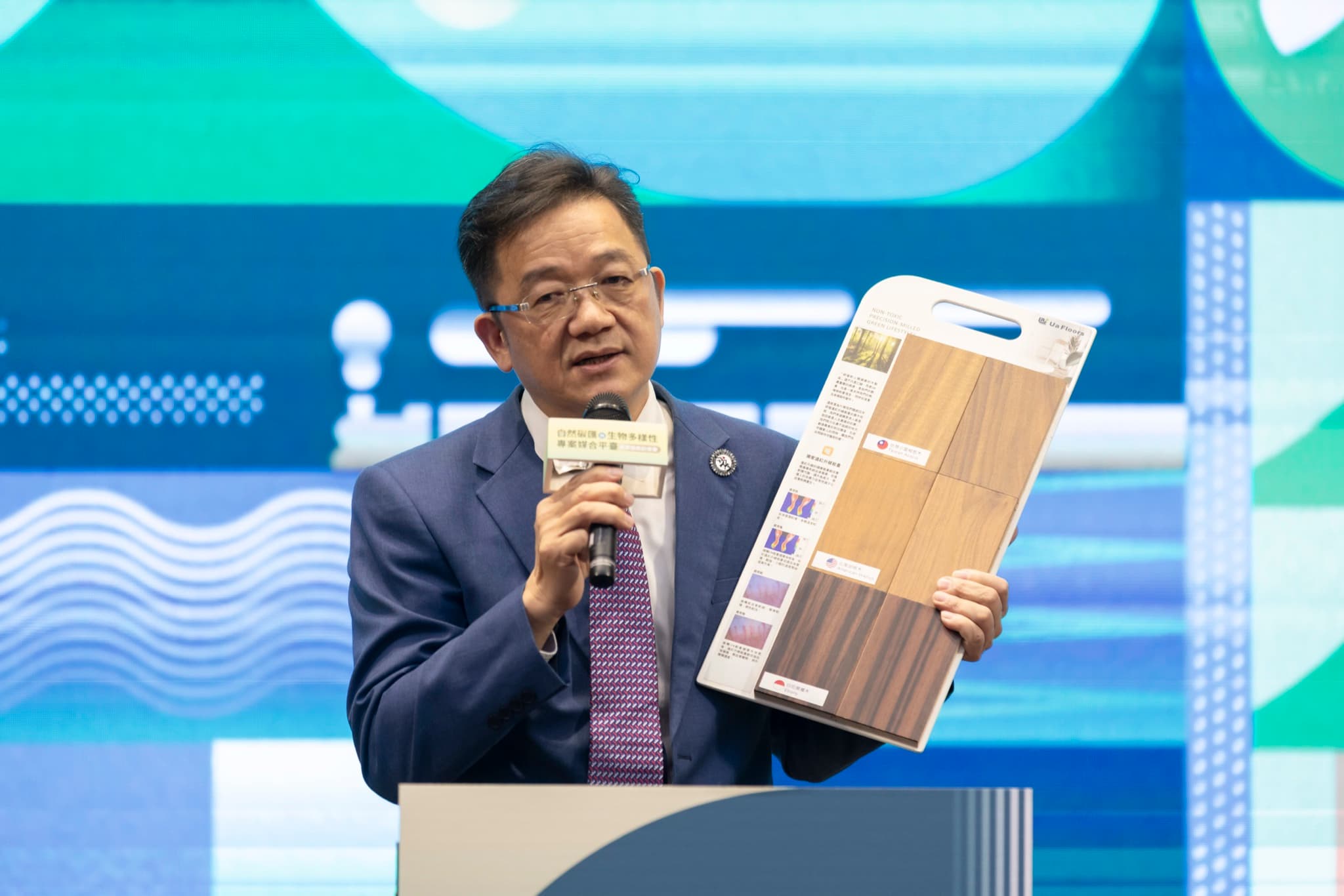 企業代表於講台上舉起木地板樣品板向台下說明,麥克風牌印有活動名稱。