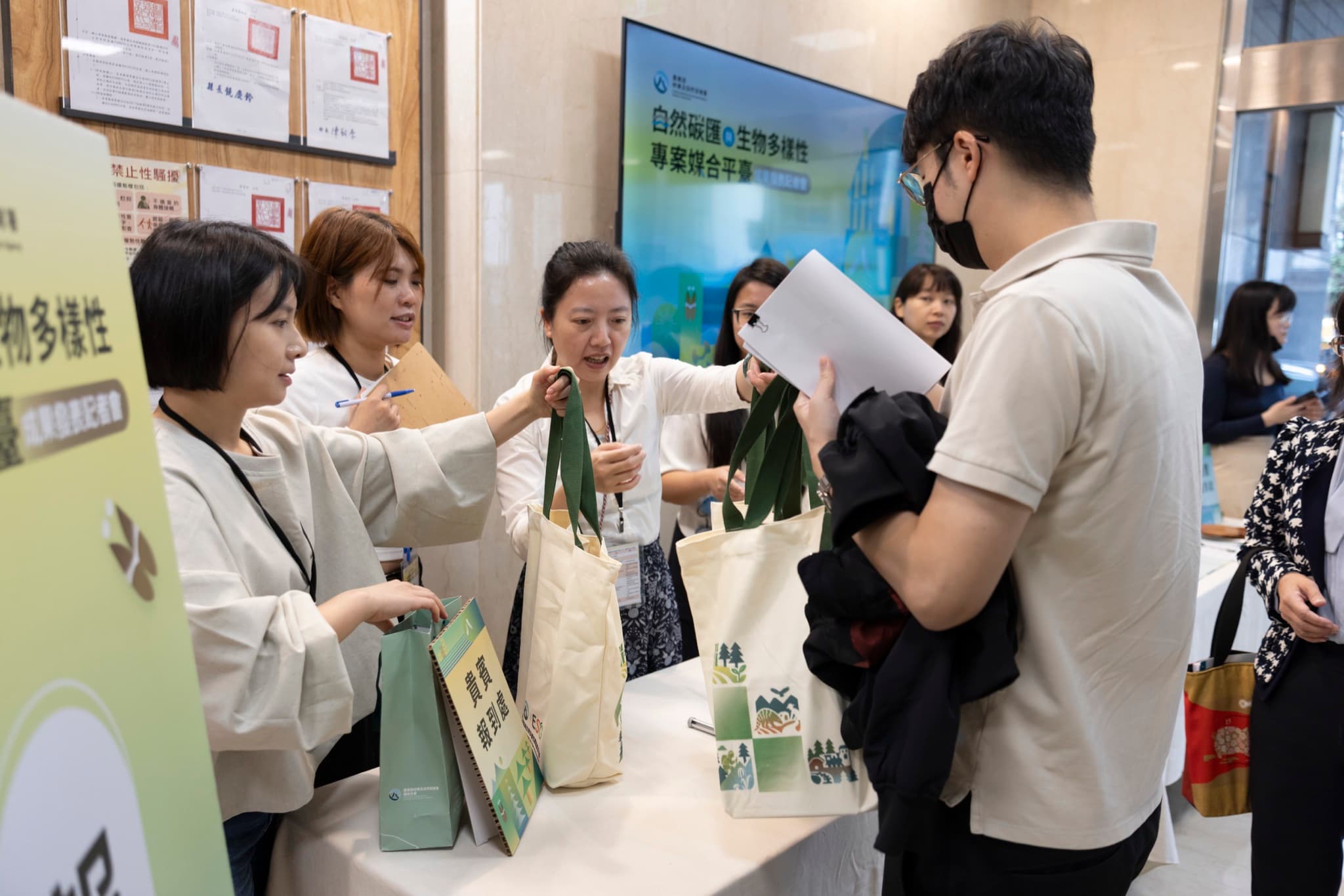 報到處工作人員向來賓遞交印有活動圖案的環保提袋,背景電視螢幕顯示記者會主視覺。