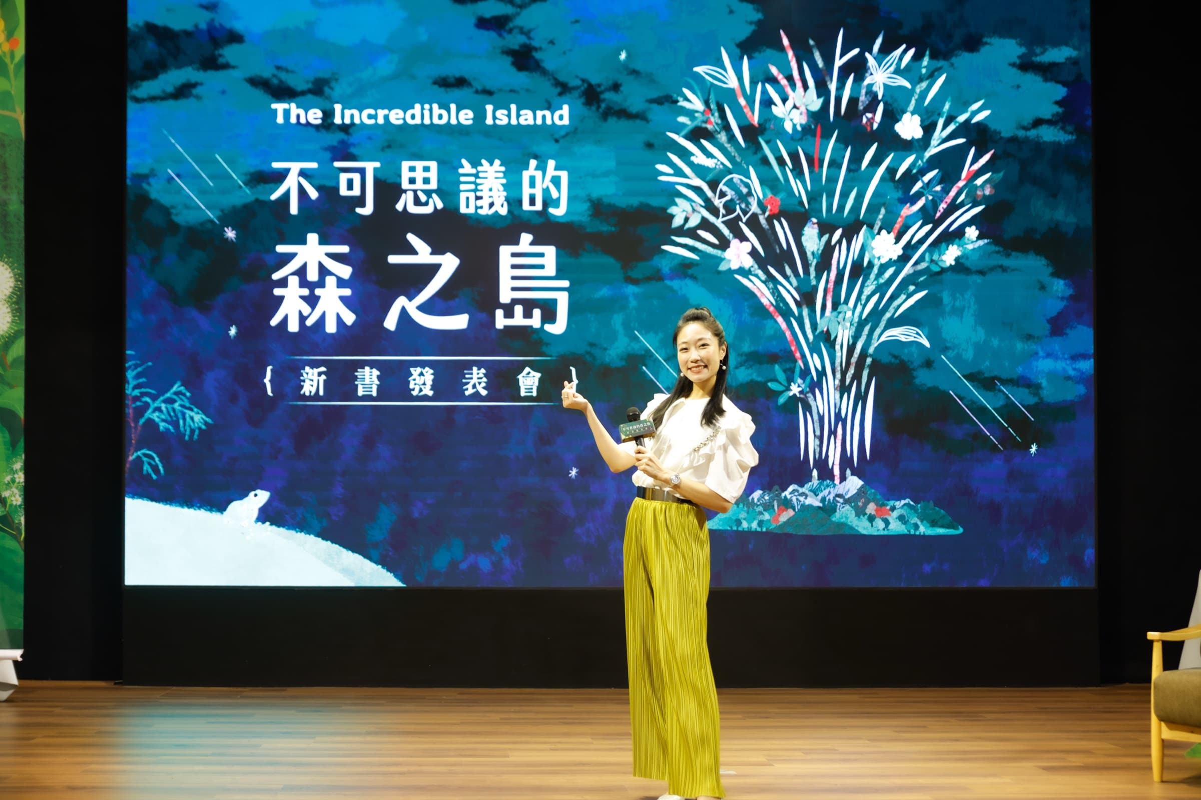 布置成森林意象的新書發表會舞台前,一位女性主持人手持麥克風,身後大螢幕為藍綠色自然插畫主視覺,一旁有座椅與植栽布置