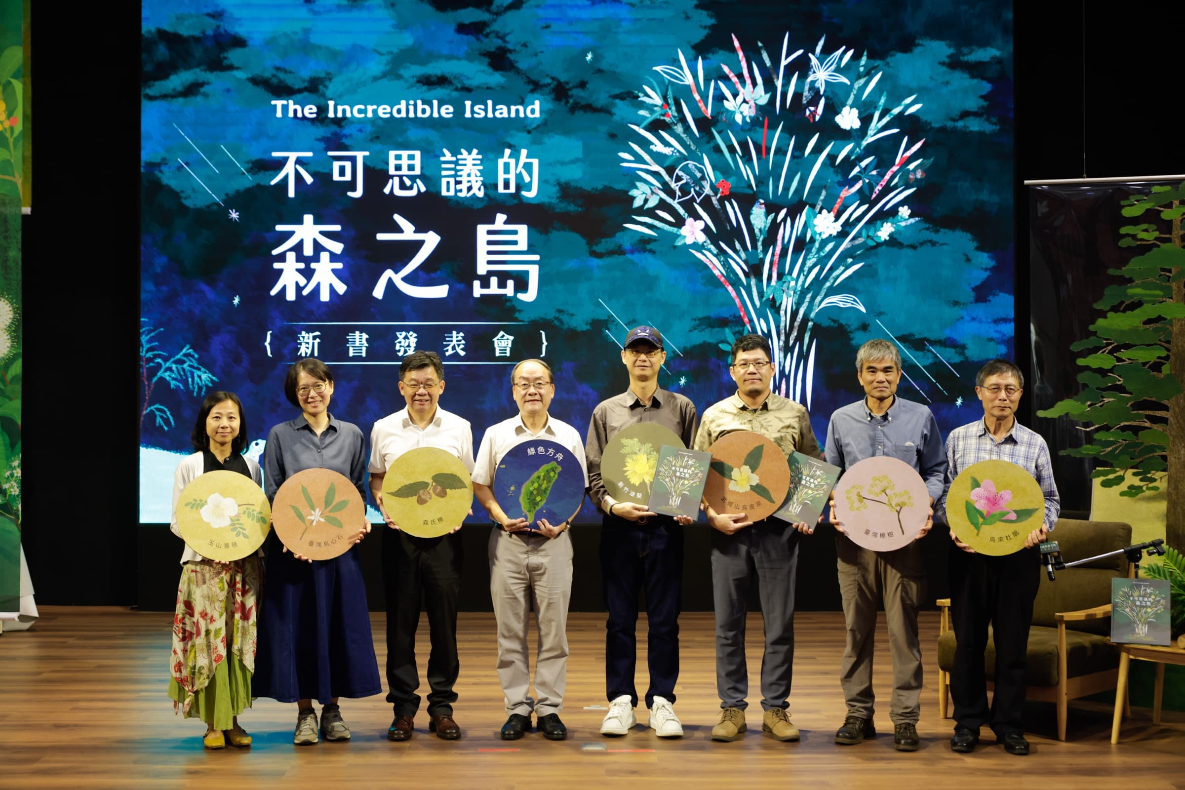 八位與會者於新書主視覺前一字排開合影,多人手持印有臺灣原生植物插畫的圓形紀念牌與繪本
