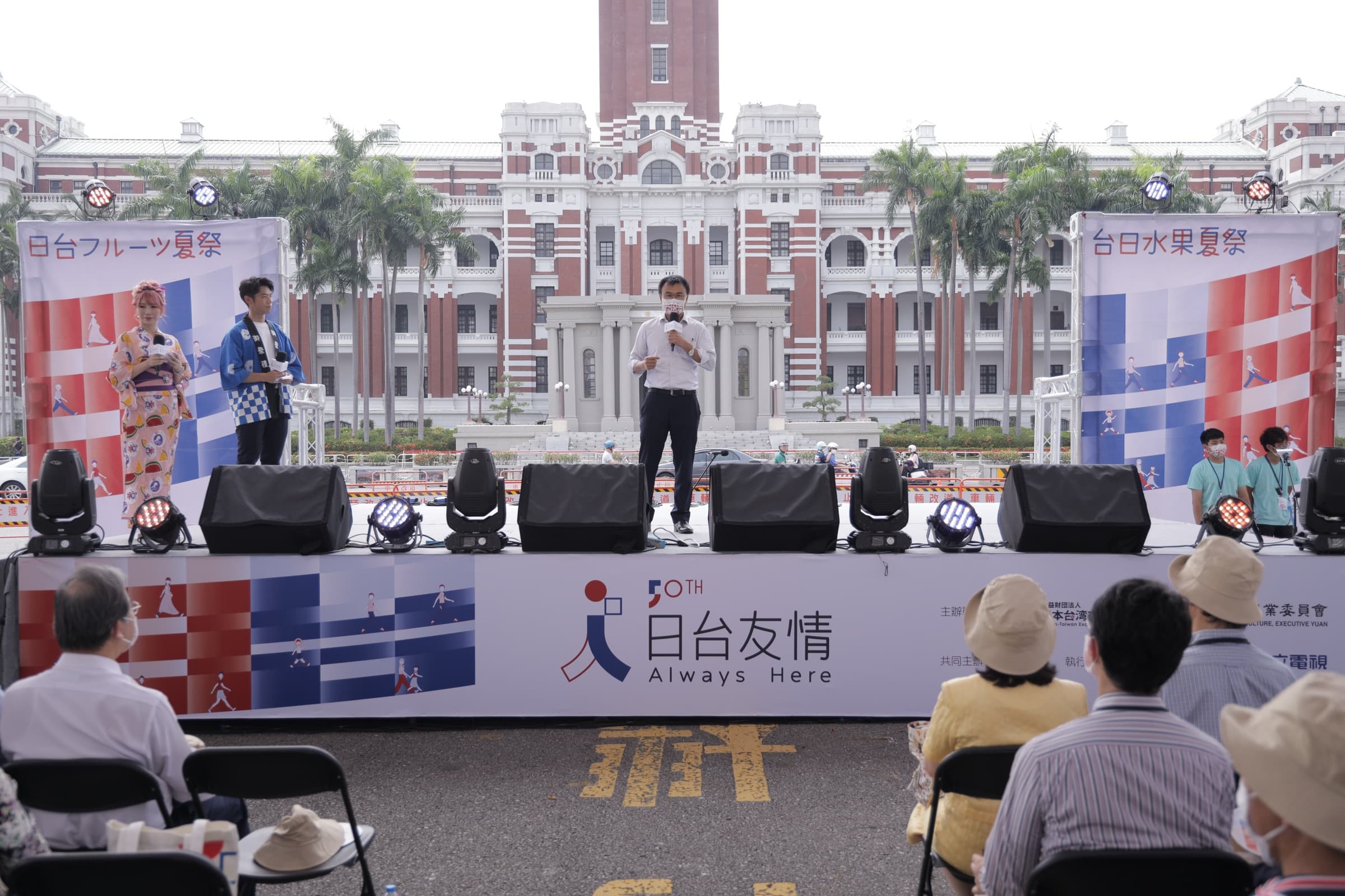 白天的台日水果夏祭舞台,一名男士於台上持麥克風致詞,左側有和服女主持與法被男主持,背景為總統府,舞台裙印有『50TH 日台友情 Always Here』與主辦識別