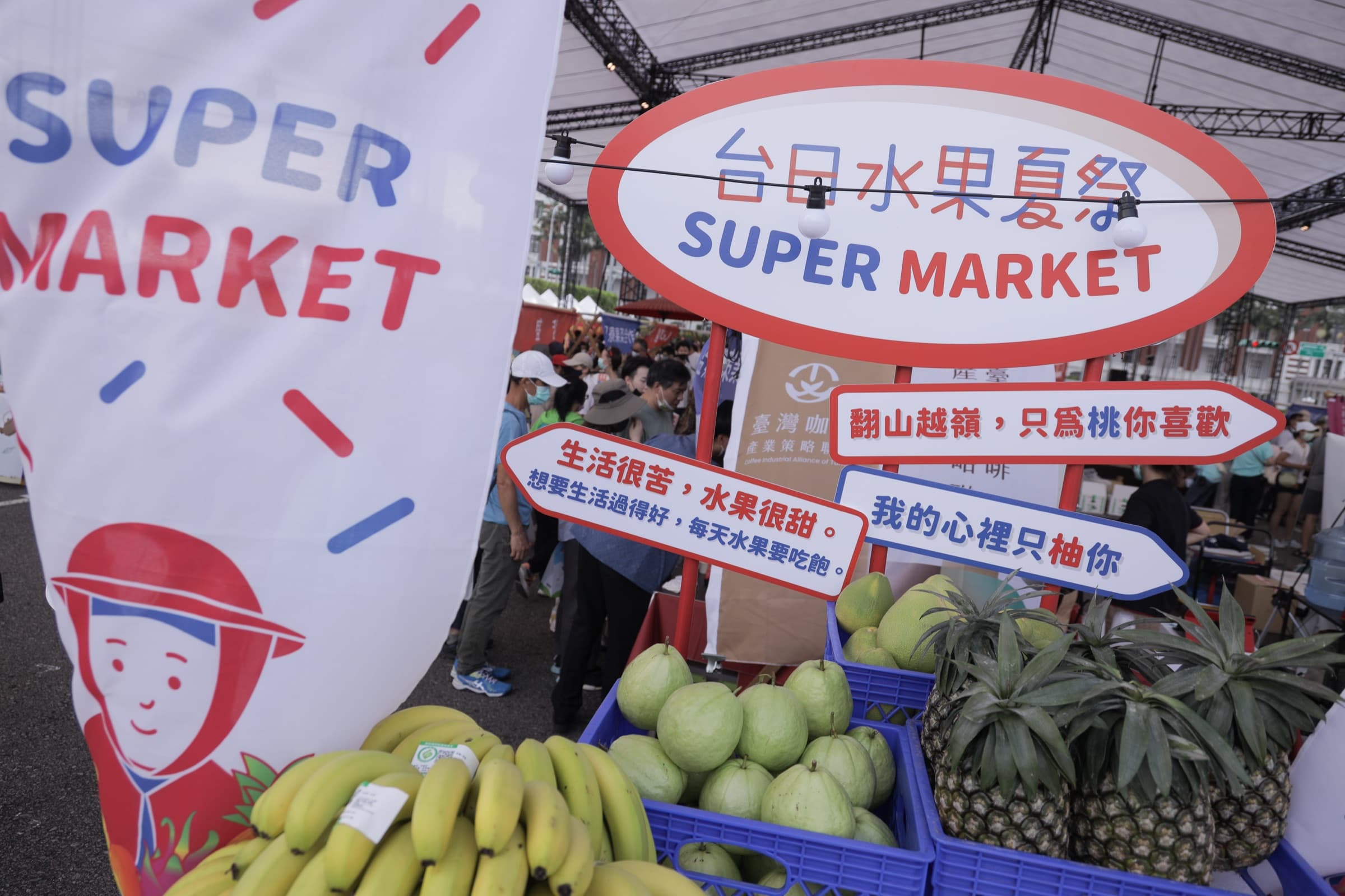 『台日水果夏祭 SUPER MARKET』圓形招牌與『翻山越嶺,只為桃你喜歡』『我的心裡只柚你』等雙關標語,下方水果攤陳列香蕉、芭樂、鳳梨、文旦