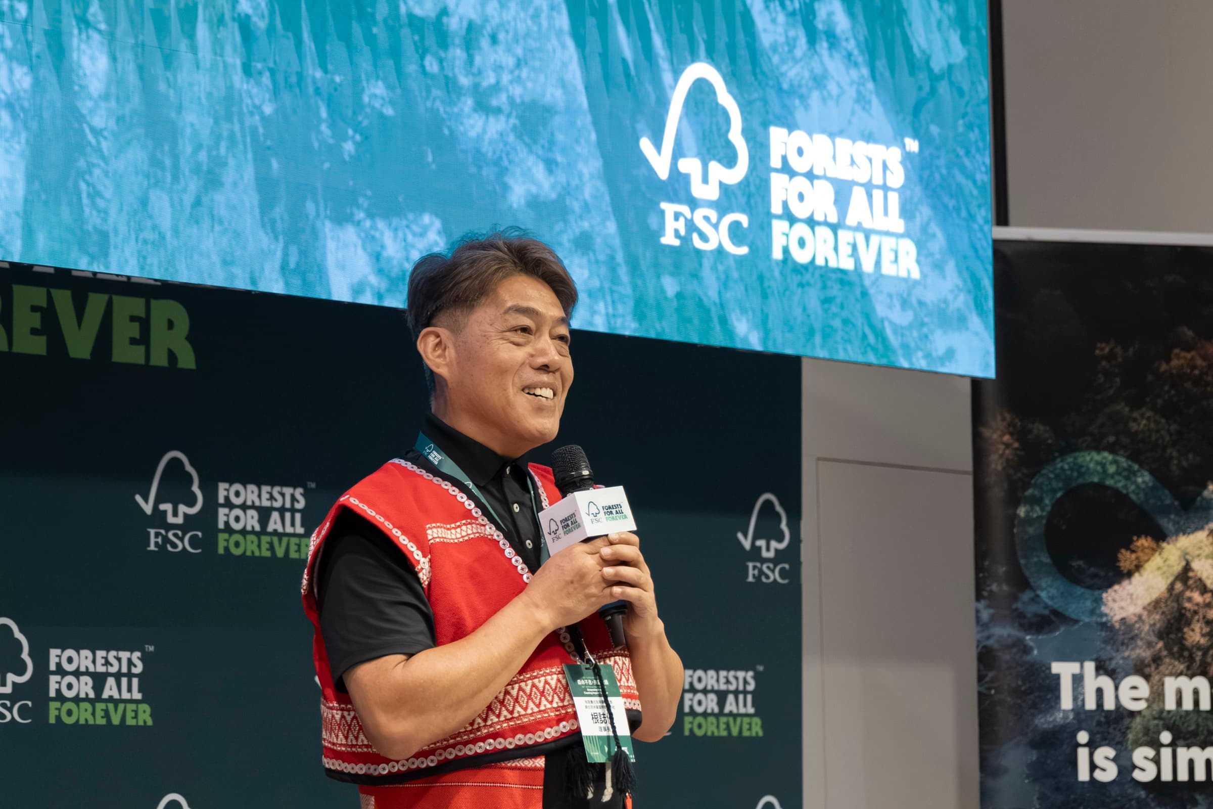 原住民族代表身著傳統紅色服飾手持麥克風,於 FSC Forests For All Forever 背板前演講