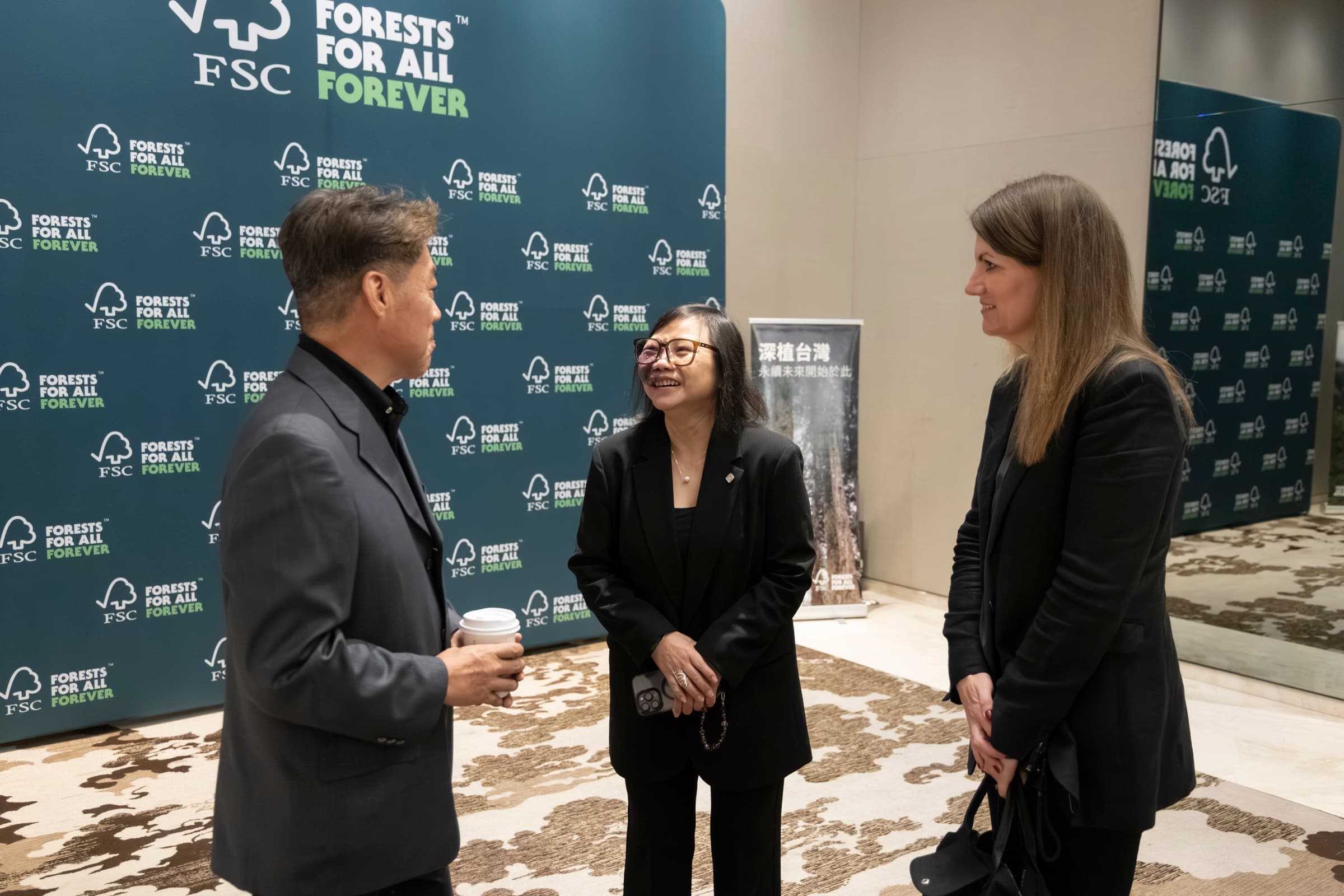 三位與會者在深色 FSC「Forests For All Forever」背板前交談,其中一人手持咖啡杯微笑
