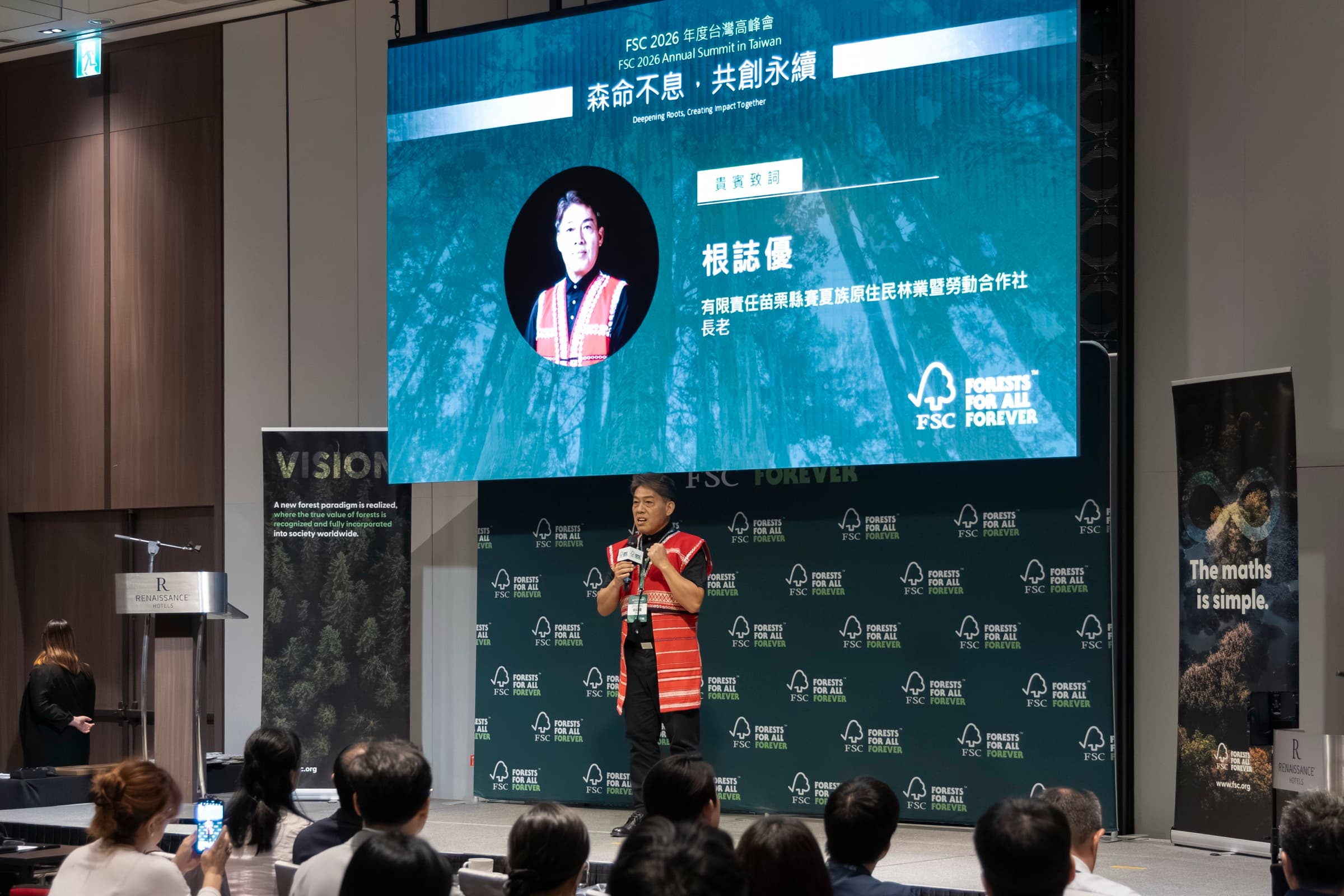 一名主辦長官站在講台前手持麥克風致詞,身後大螢幕顯示其姓名與職稱,背板為 FSC 主視覺