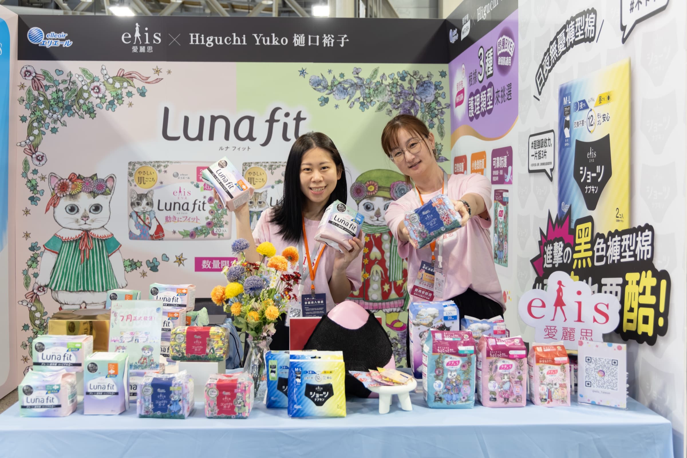 女子月月有特展參展品牌攤位,兩位工作人員手持女性生理用品向鏡頭展示並微笑,攤位牆面滿是品牌主視覺與商品陳列