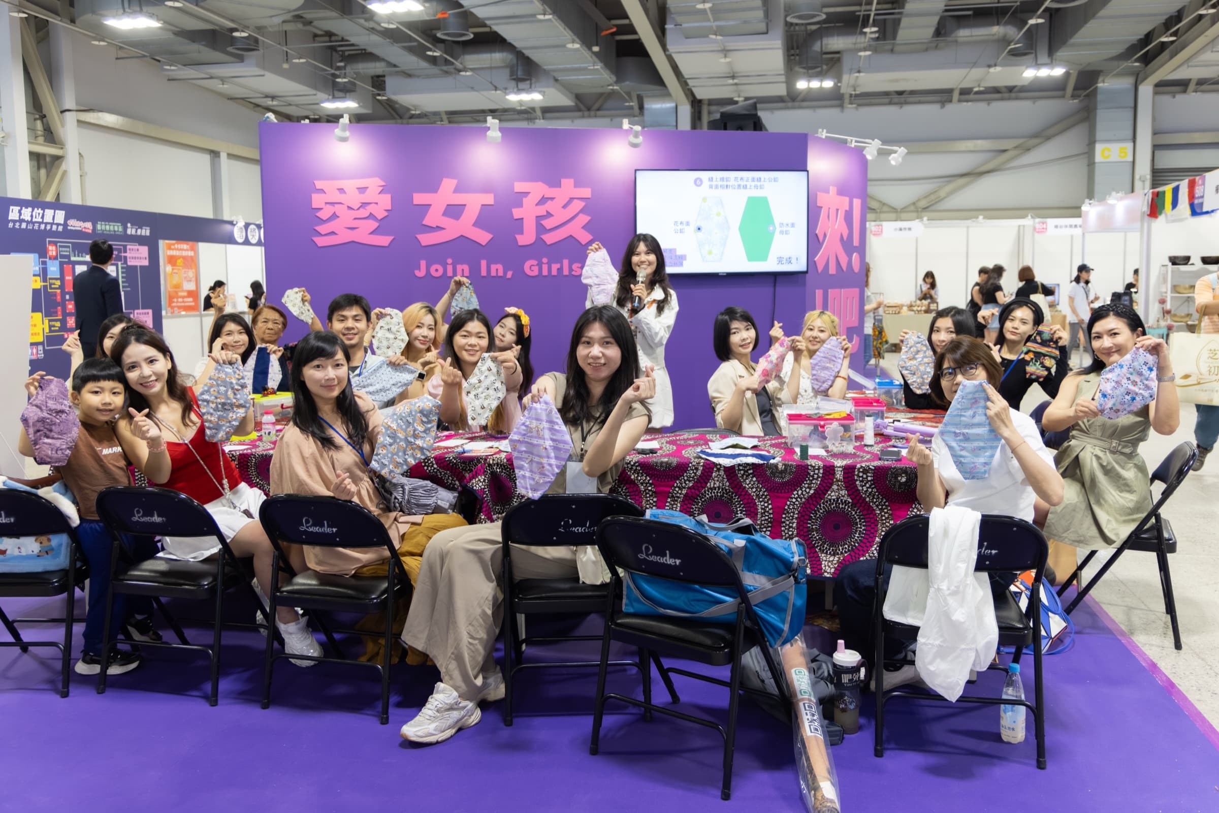 女子月月有特展『愛女孩 Join In, Girls』公益展區,整桌學員舉起自己手縫完成的布衛生棉比愛心,講師持麥克風帶領,螢幕顯示縫製步驟