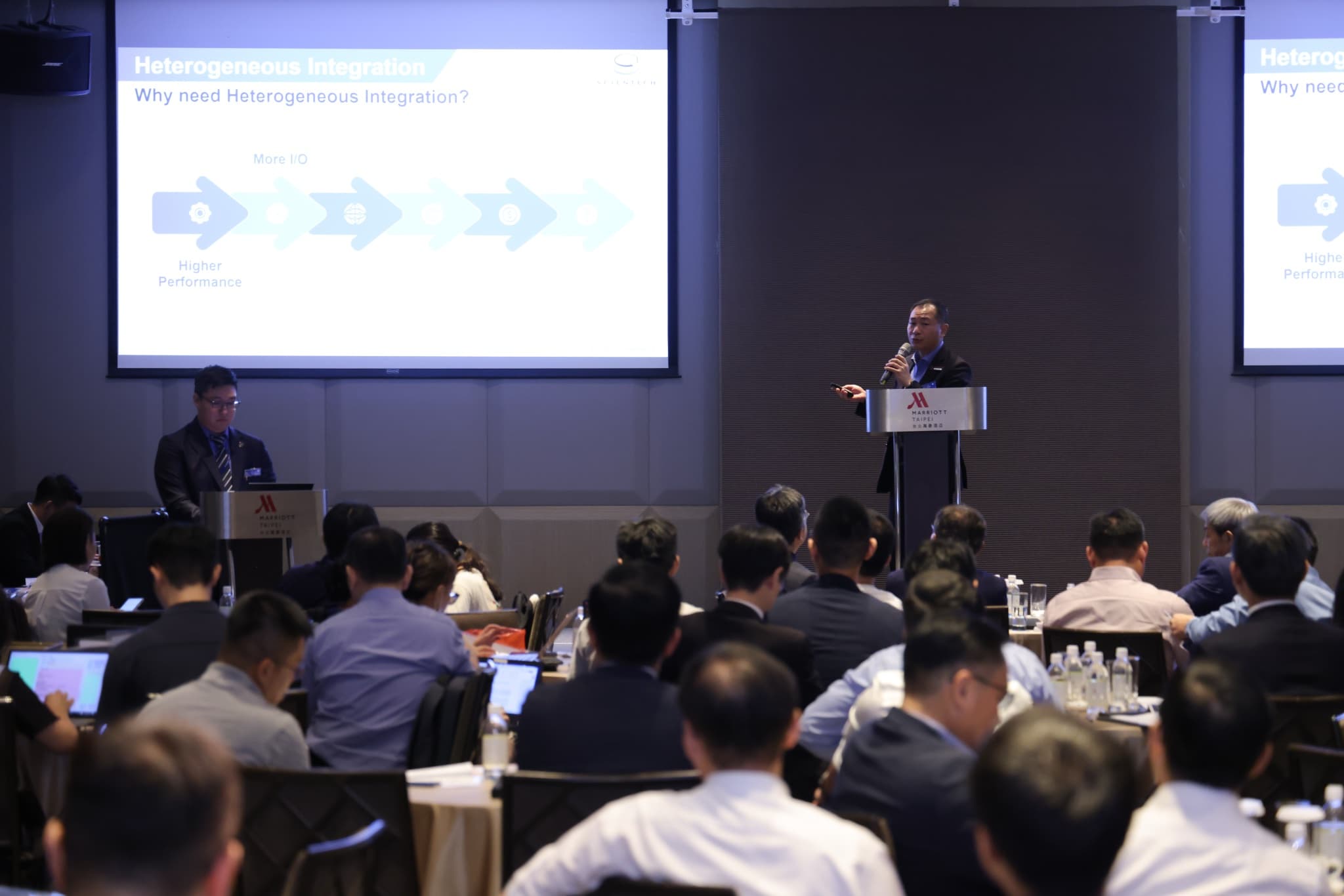 雙投影幕同步顯示 Heterogeneous Integration 簡報,講者站在講台旁,台下圓桌坐滿與會者