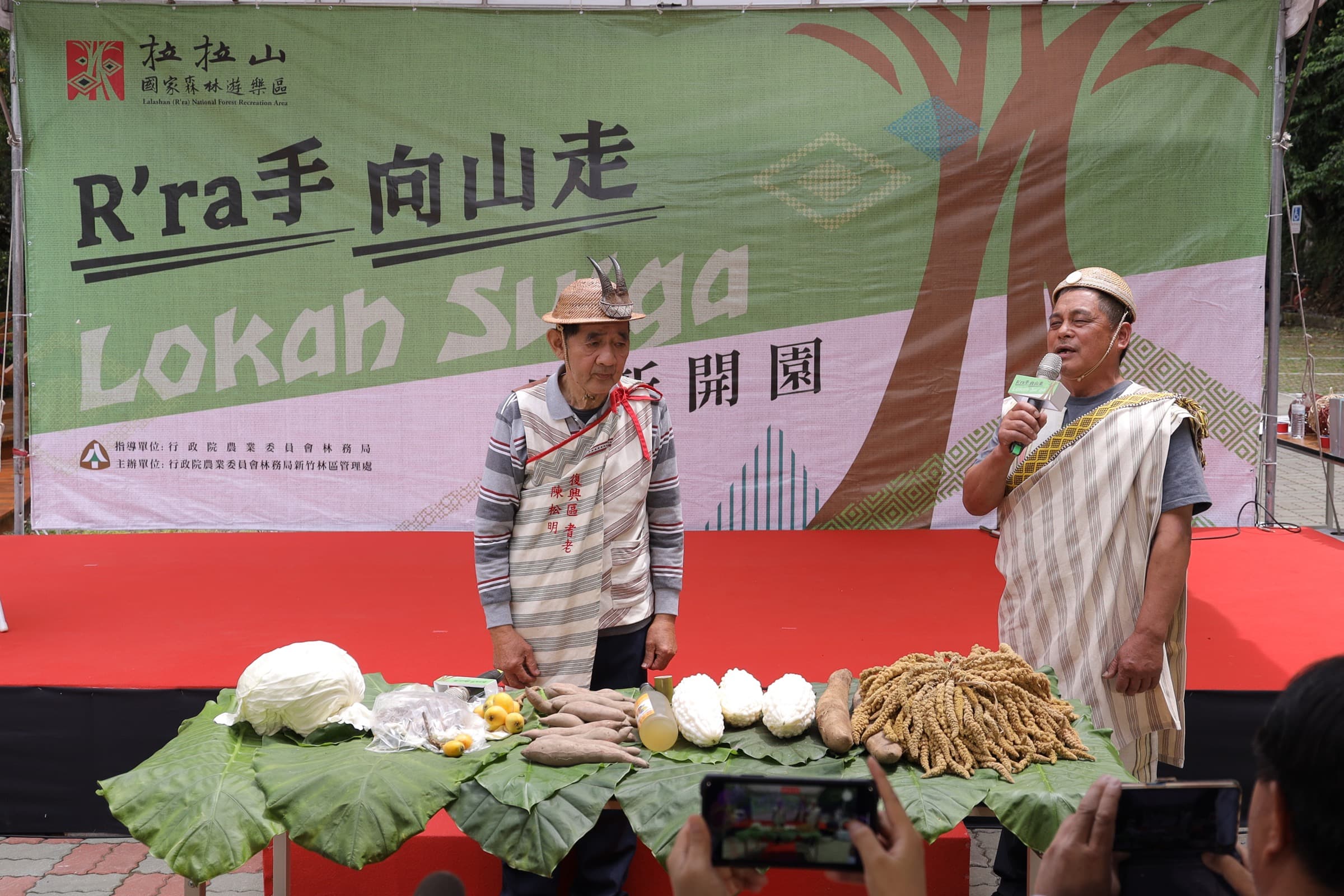 舞台上原民服飾男性持麥克風於祭品桌前致詞