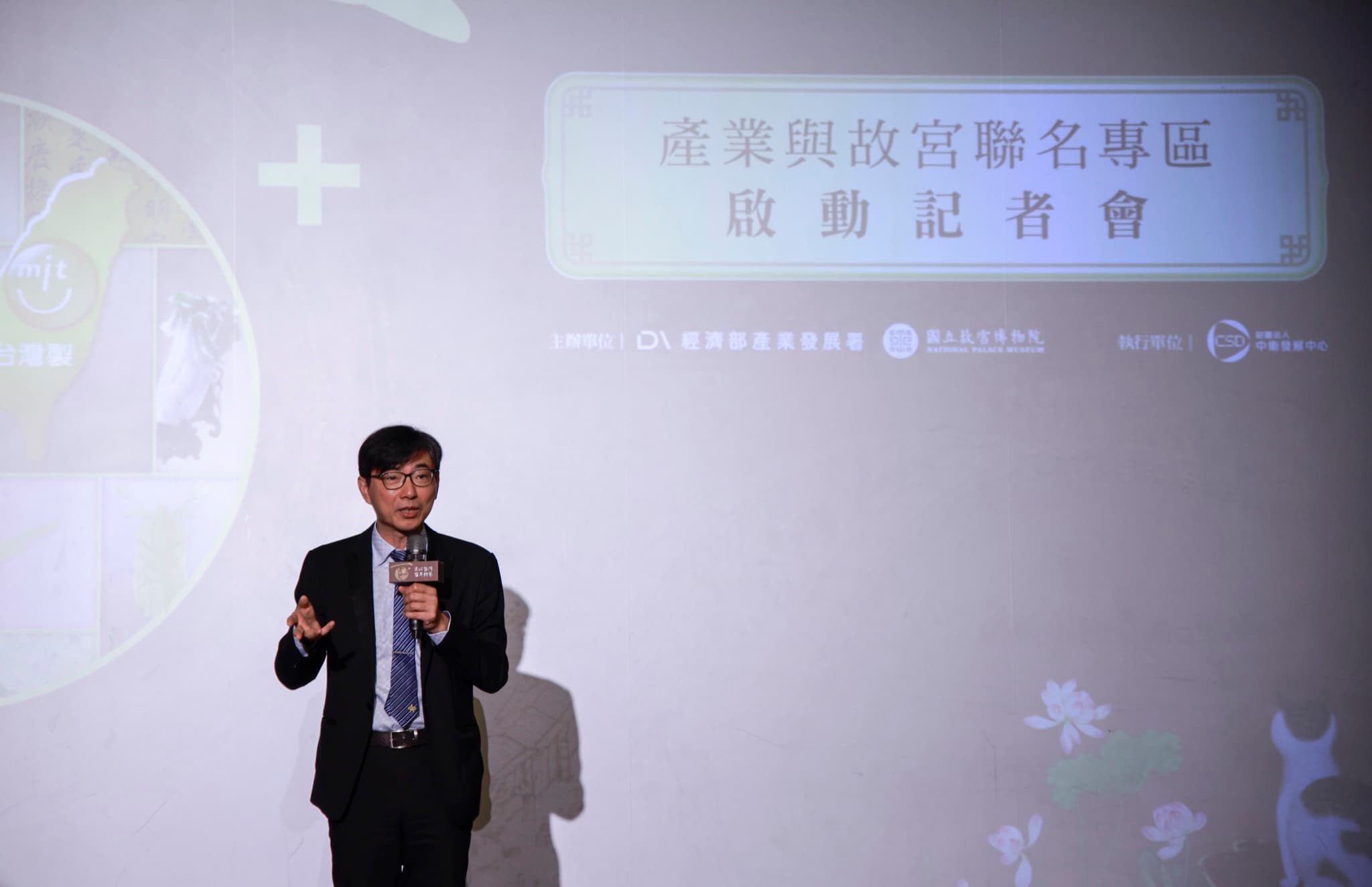 講者持印有活動主視覺的麥克風致詞,身後投影幕列出主辦單位經濟部產業發展署與國立故宮博物院