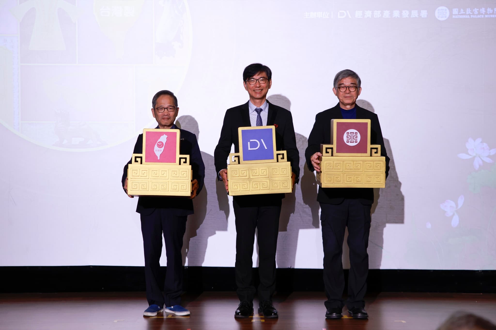 三位代表各持印有台灣MIT微笑標章、經濟部產業發展署、故宮院徽的金色寶盒道具並排合影,螢幕上方標示主辦單位字樣