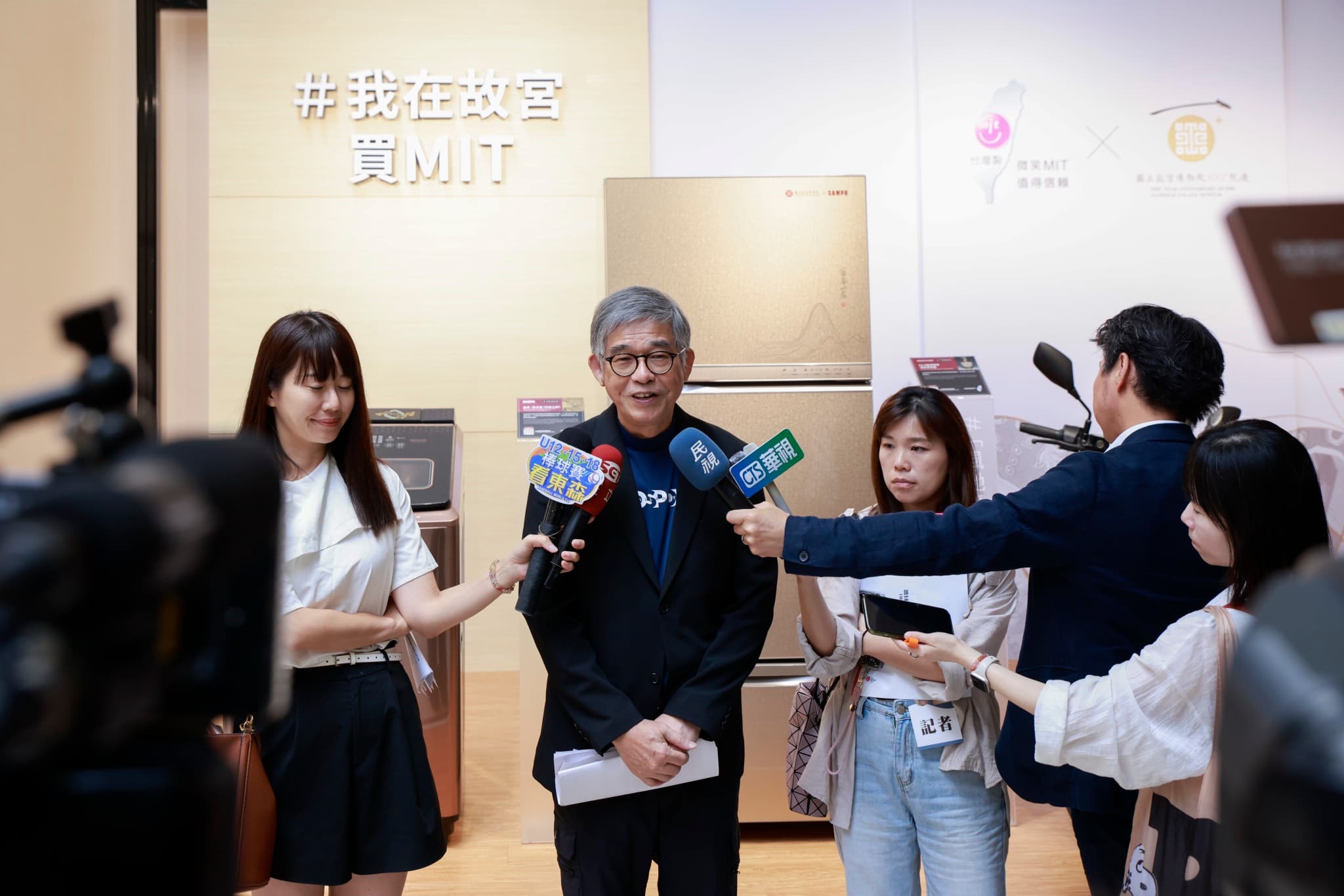 貴賓在「#我在故宮 買MIT」標語牆前接受多家電視台記者持麥克風聯訪