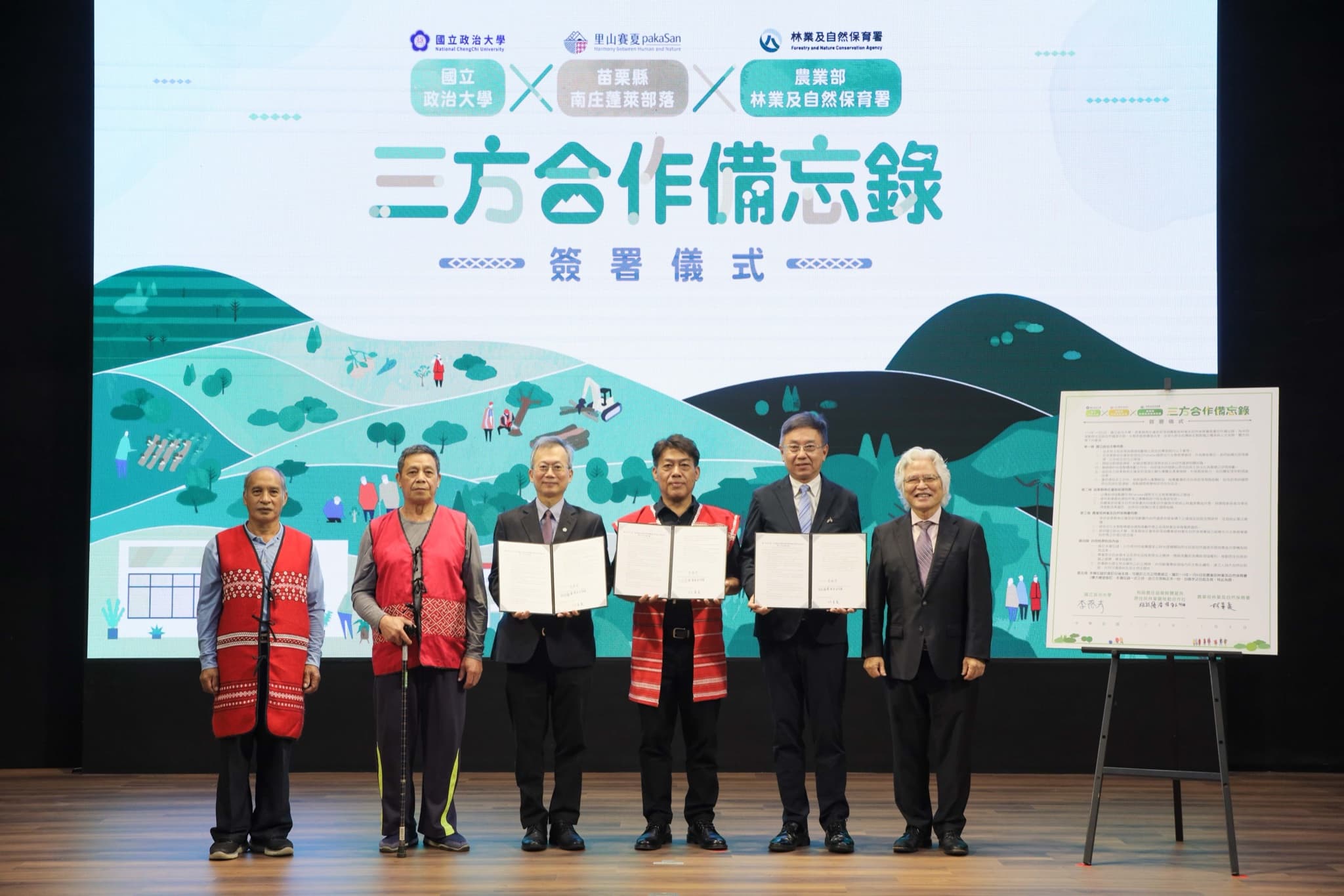 六位代表並排展示三份打開的簽署文件,中央代表著賽夏族紅色族服,右側畫架立著簽妥的備忘錄看板