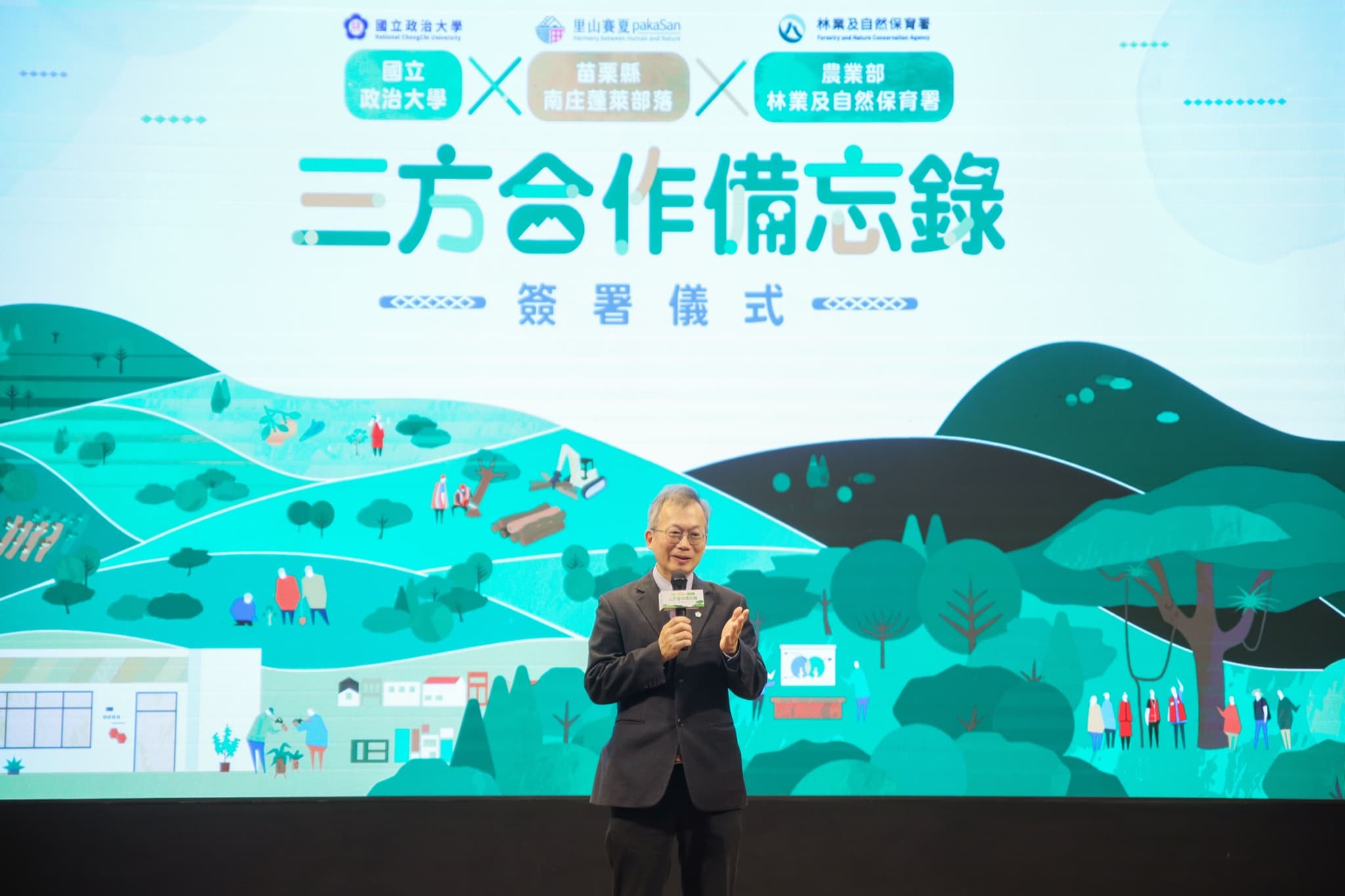 一位西裝男士站在三方合作備忘錄簽署儀式背板前持麥克風致詞,背板列出國立政治大學、苗栗縣南庄蓬萊部落、農業部林業及自然保育署