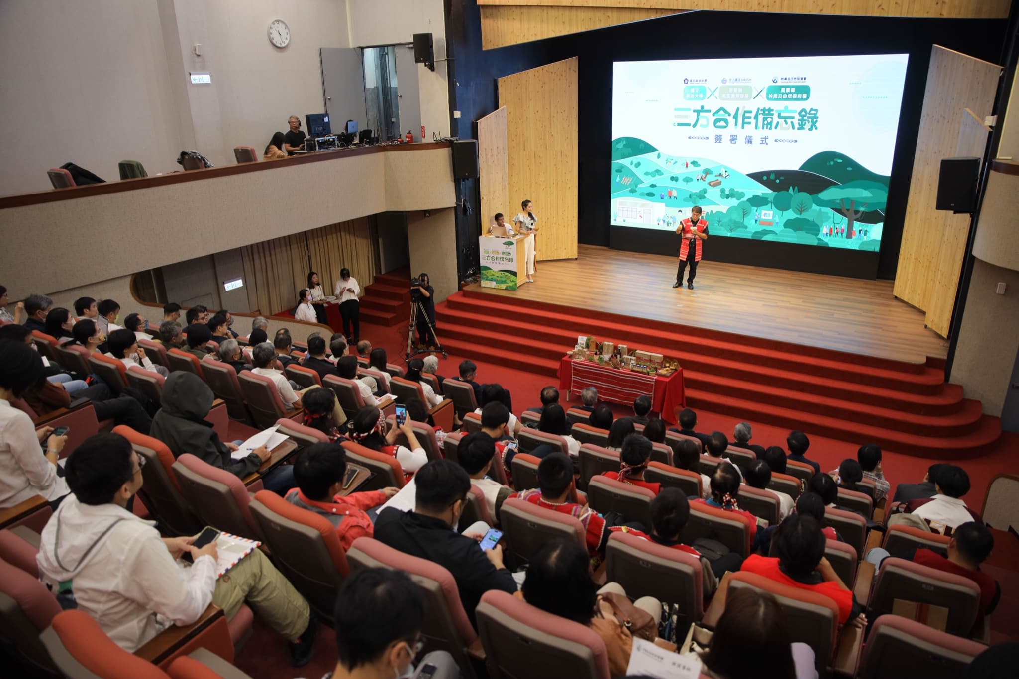 由觀眾席後方俯瞰禮堂全景,滿座觀眾面向舞台,台上部落代表致詞,背板與農產展示桌入鏡