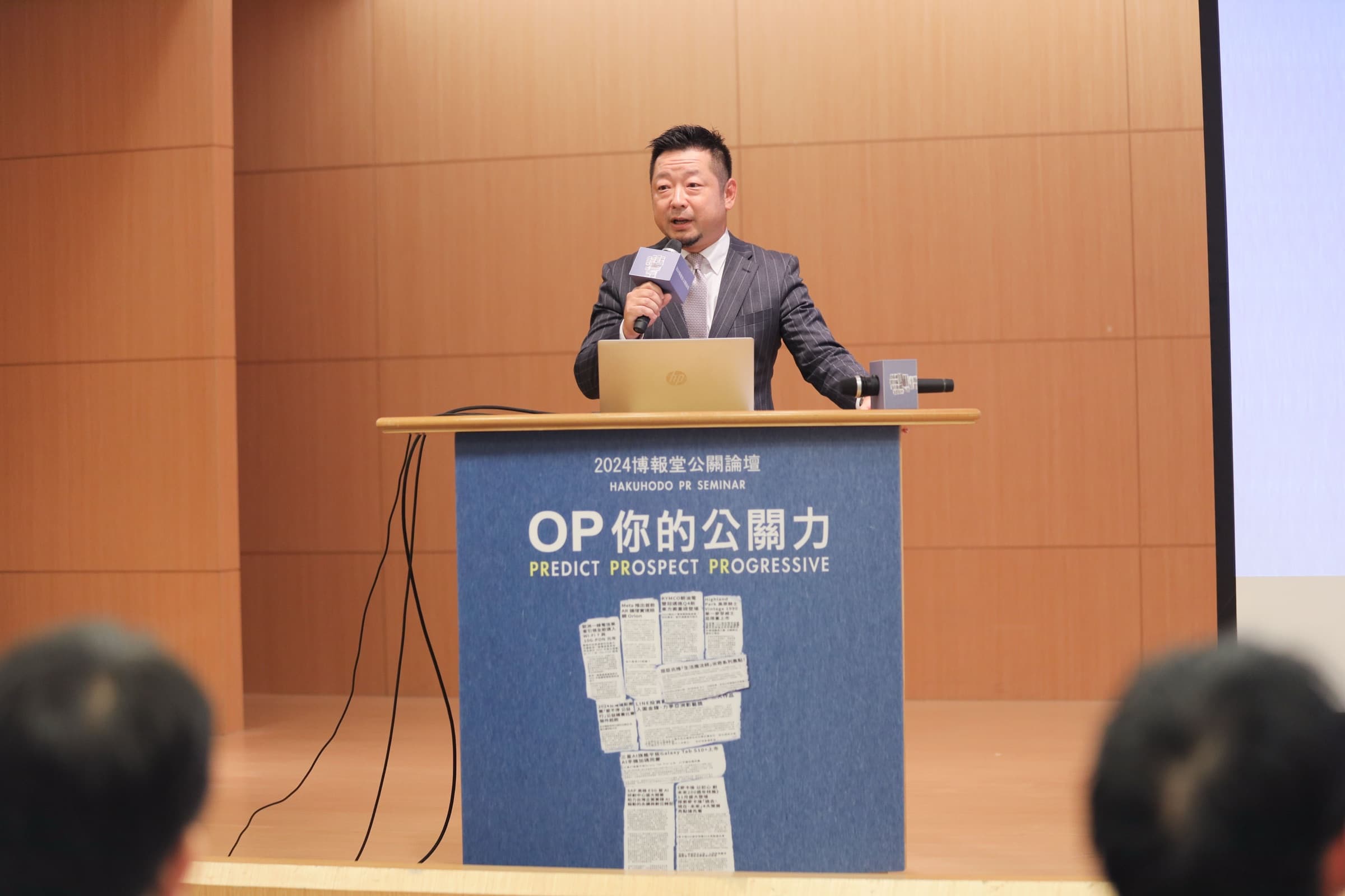 一位男性講者於印有『2024 博報堂公關論壇 OP 你的公關力』主視覺的講台後,手持麥克風演講