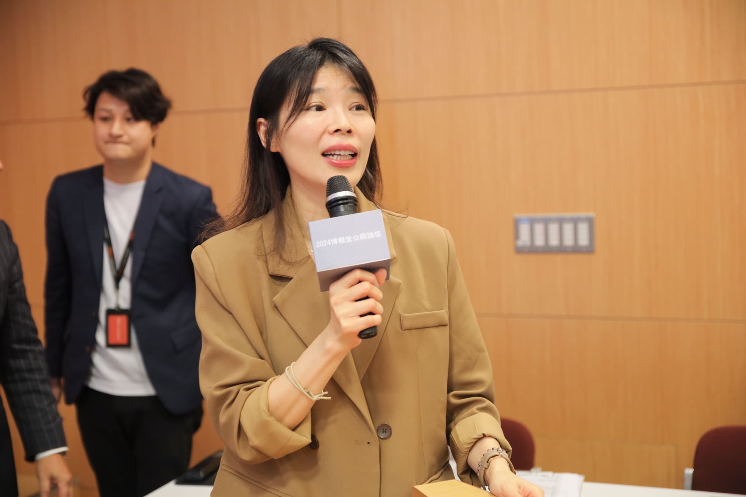 一位身著駝色西裝的女性講者手持麥克風、面帶笑容分享,麥克風牌寫著『2024博報堂公關論壇』