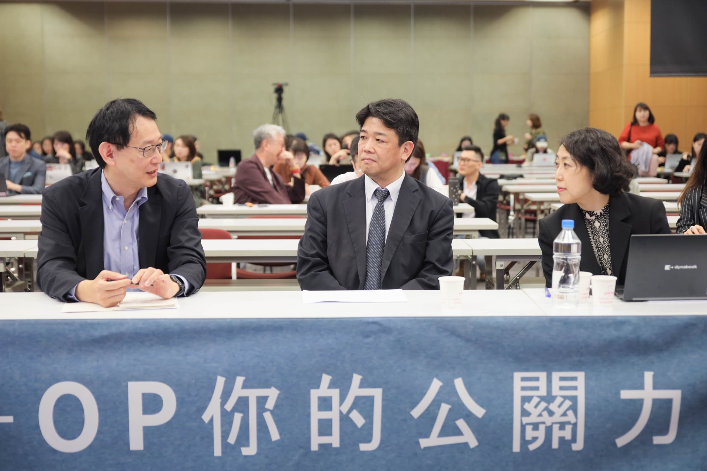 三位與談人於鋪著『OP 你的公關力』桌布的長桌後就座交流,背景為坐著聽講的觀眾席