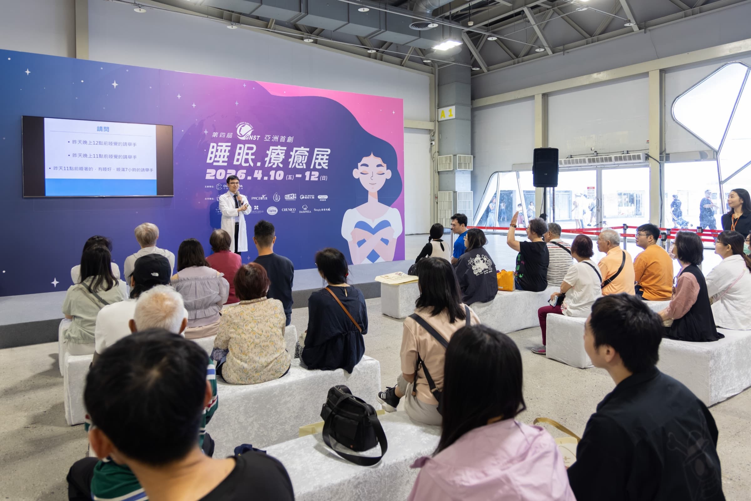 白袍睡眠專科醫師於展區講台持麥克風開講,大螢幕顯示睡眠自我檢視題目,台下觀眾就座聆聽,背板為睡眠療癒展主視覺