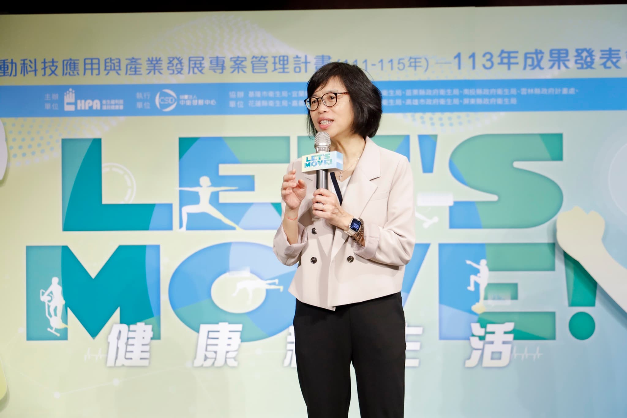 國民健康署官員持 Let's Move 麥克風牌致詞,背板可見計畫全銜與主辦單位衛生福利部國民健康署標誌