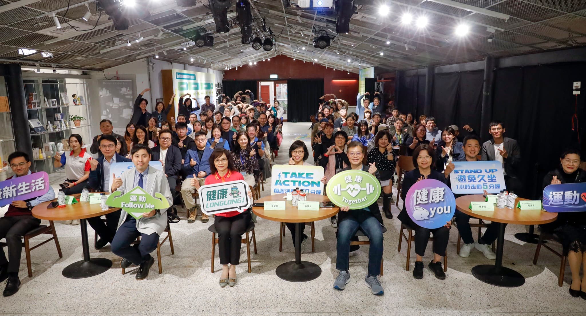 全場約百人大合照,前排貴賓手持「TAKE ACTION」「健康新生活」「運動 Follow Me」等標語板,場地縱深完整入鏡
