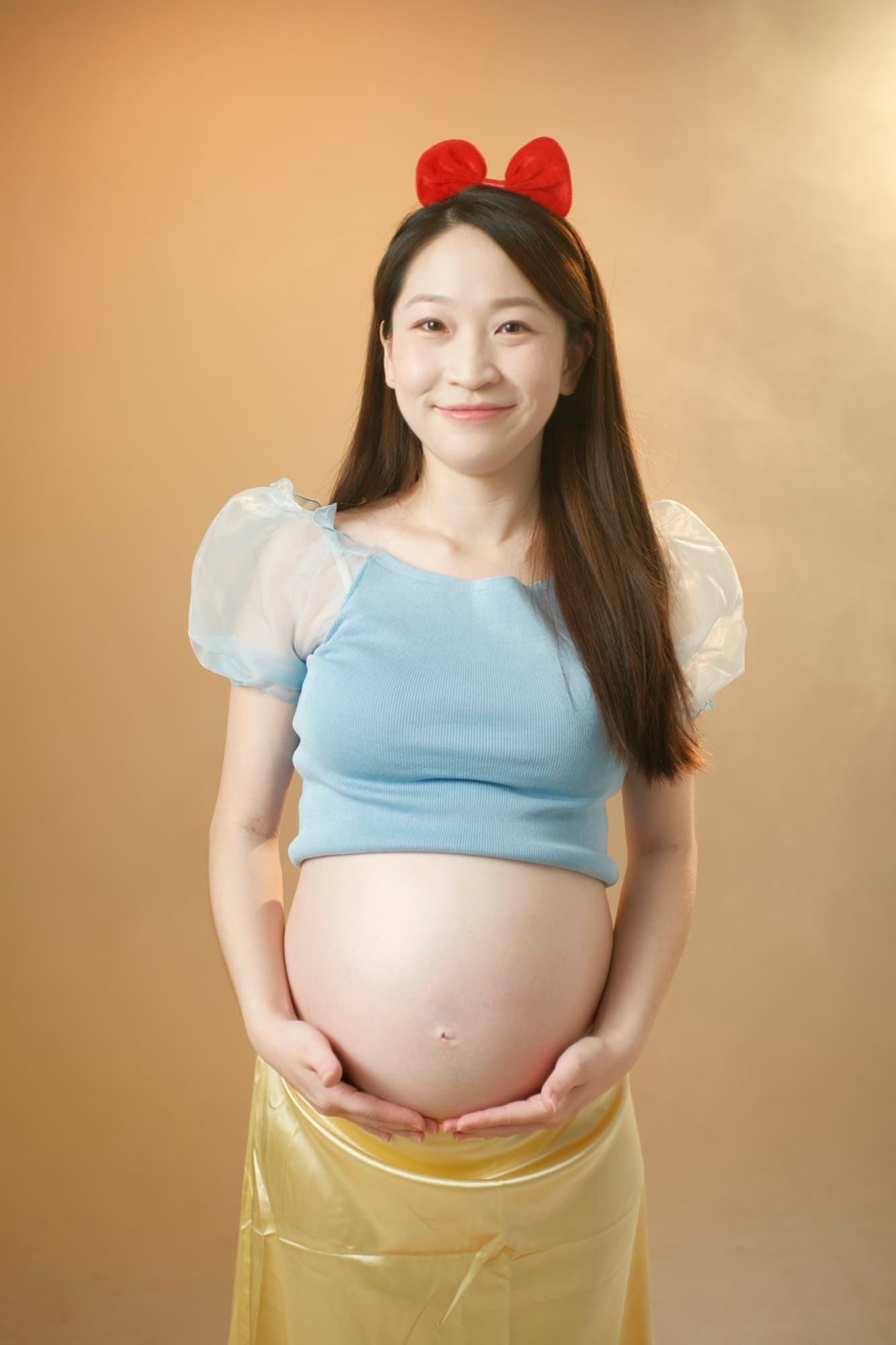 藍上衣紅蝴蝶結孕婦雙手托腹、正面微笑的七分身像
