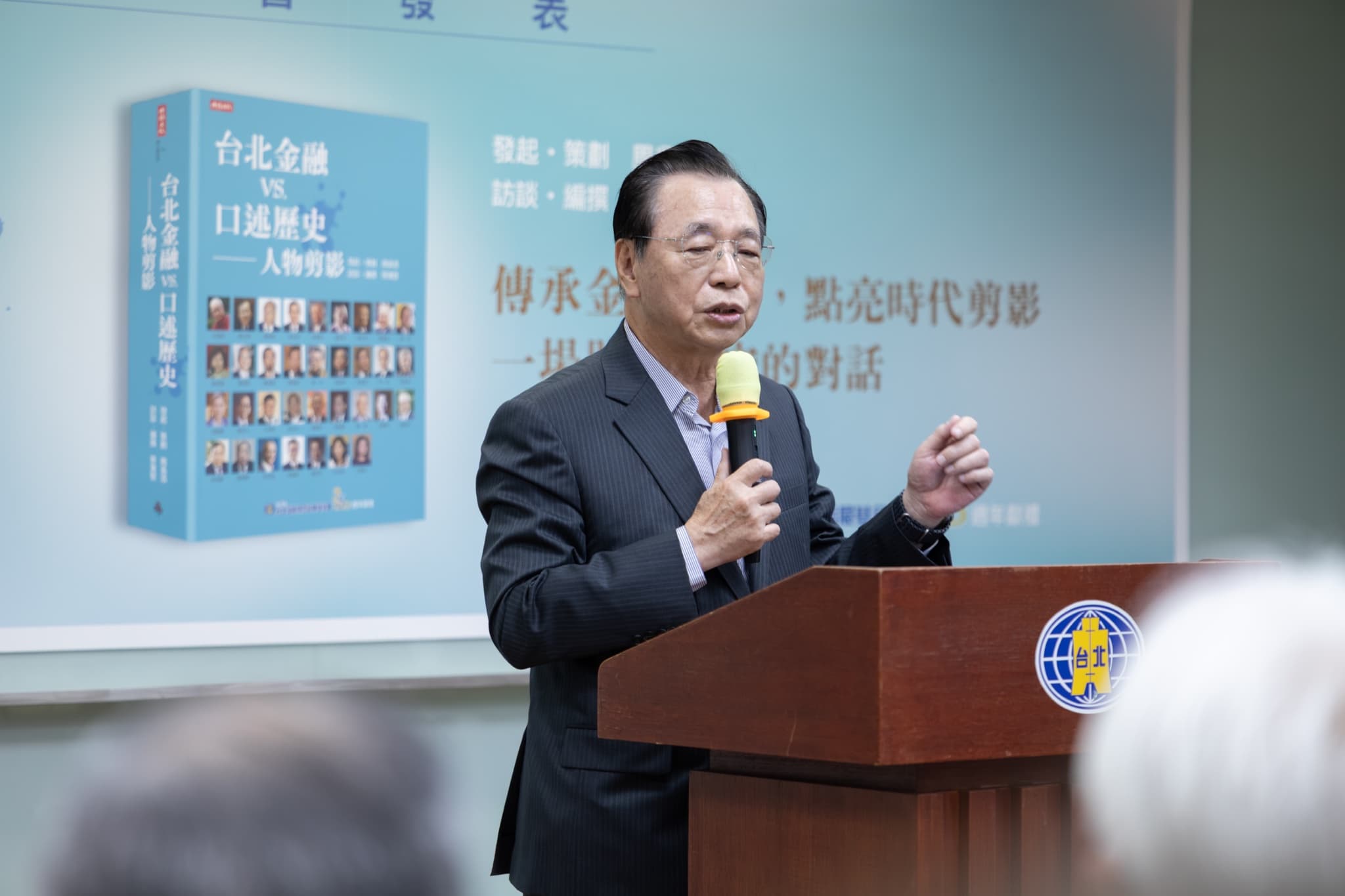 貴賓於木質講台手持麥克風致詞,身後背板為新書書封與「傳承金融薪火,點亮時代剪影」標語