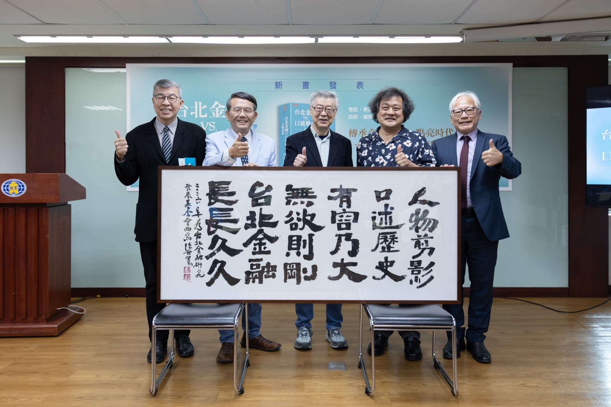五位貴賓在背板前與「人物剪影 口述歷史 有容乃大 無欲則剛 台北金融 長長久久」書法作品合影比讚
