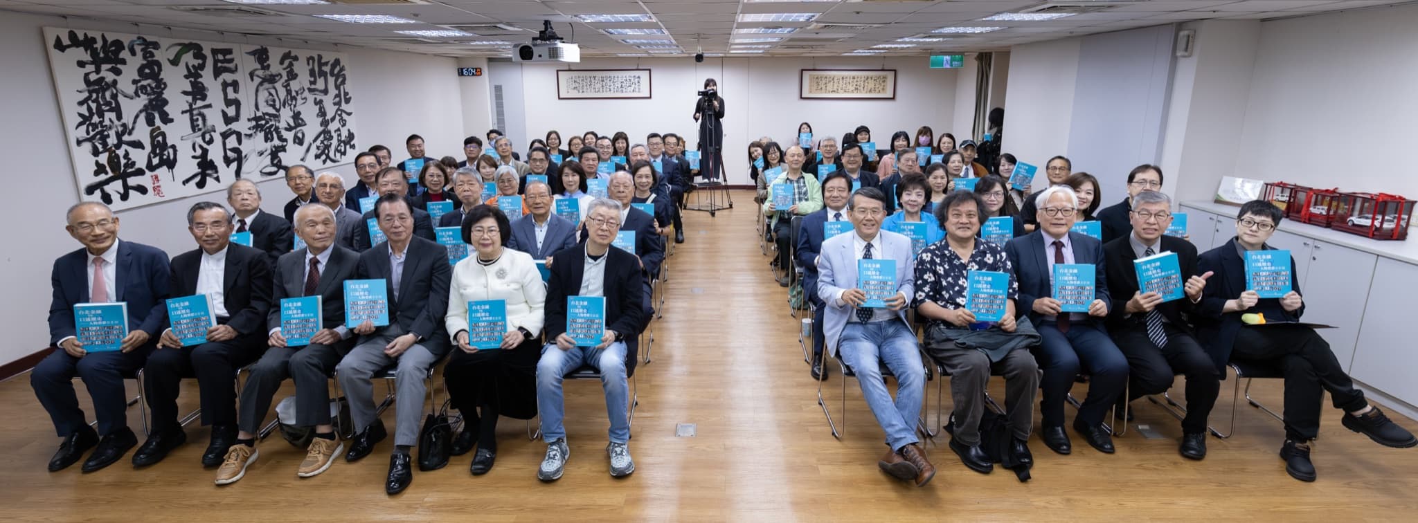 全體與會來賓手持藍色新書於會場大合照,前排資深貴賓就座、後排起立,超寬幅構圖