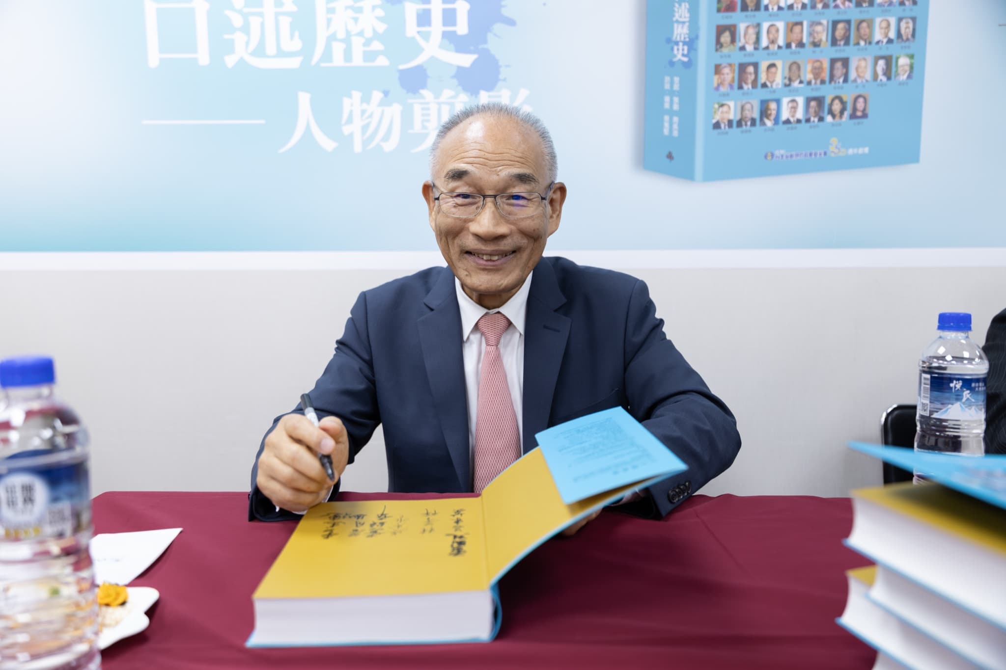 貴賓於酒紅桌巾簽書桌前微笑題字,新書攤開至已集滿簽名的黃色蝴蝶頁