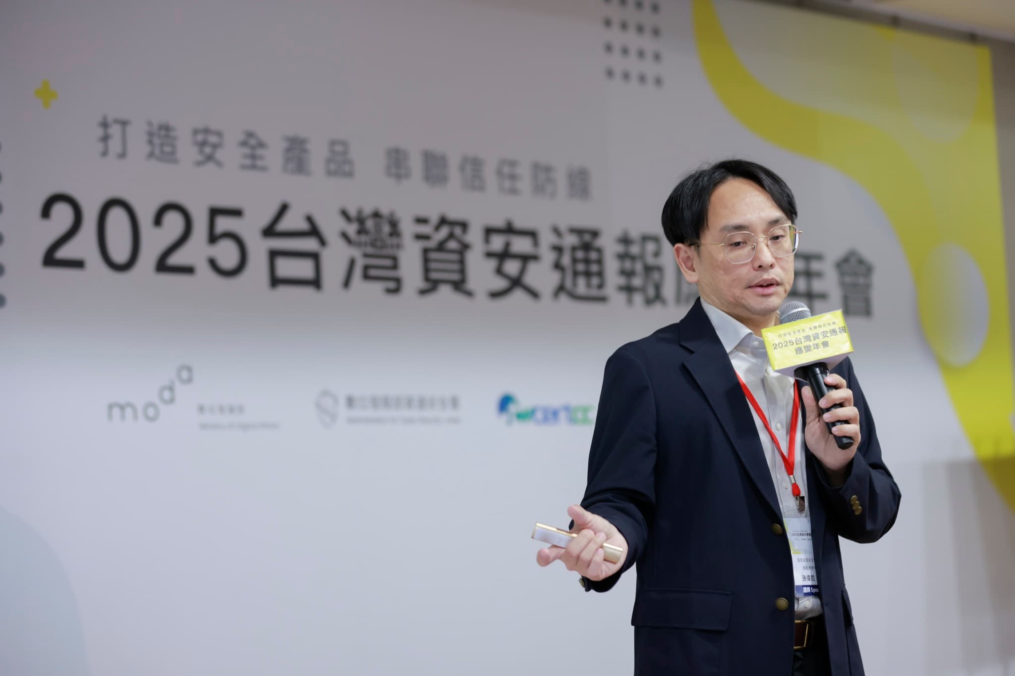 講者站於舞台中央手持印有年會名稱的麥克風牌演講,背板四個主辦單位logo清晰可見