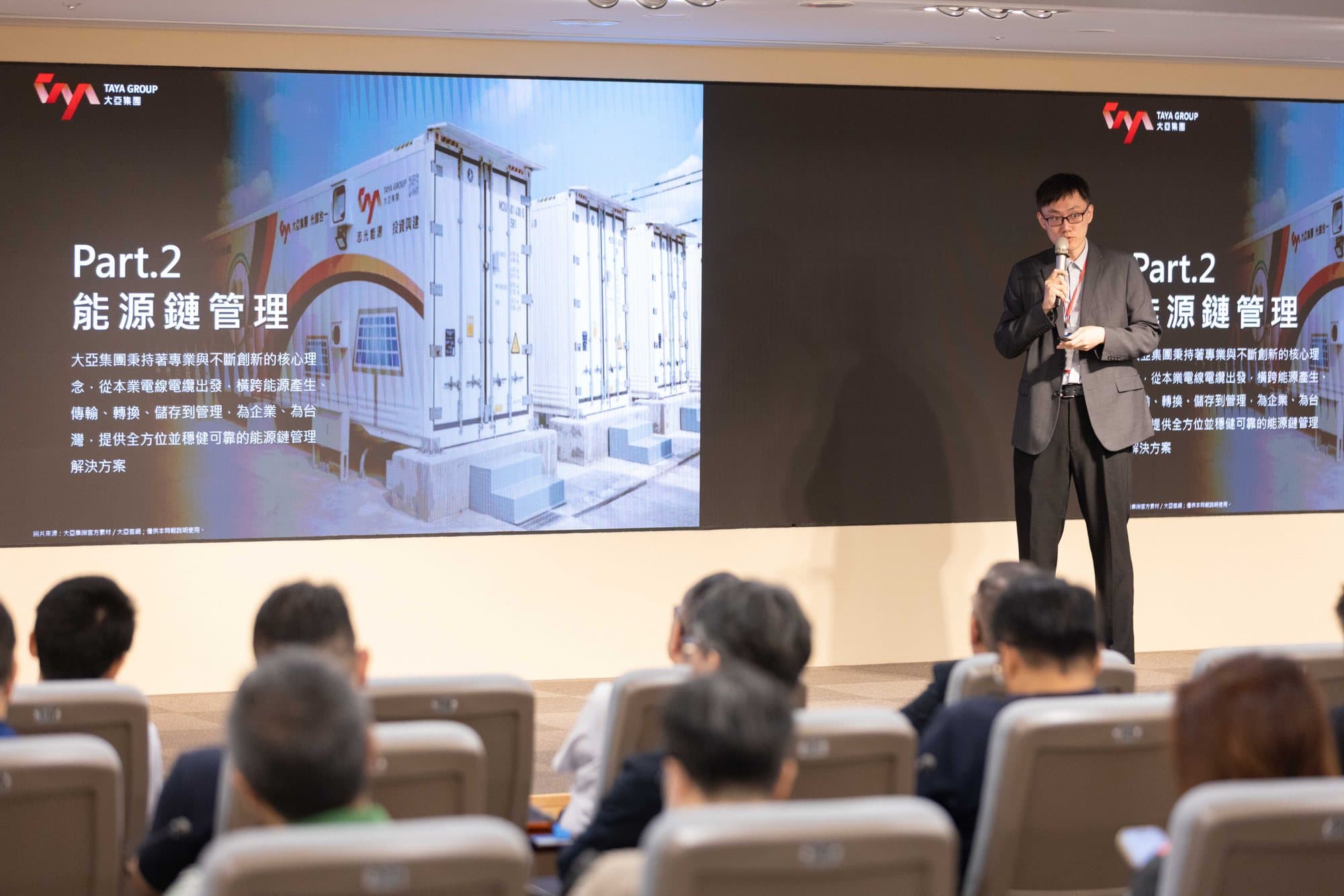 講者站在舞台上演講,身後兩面 LED 螢幕同步顯示簡報,前景是觀眾席後腦